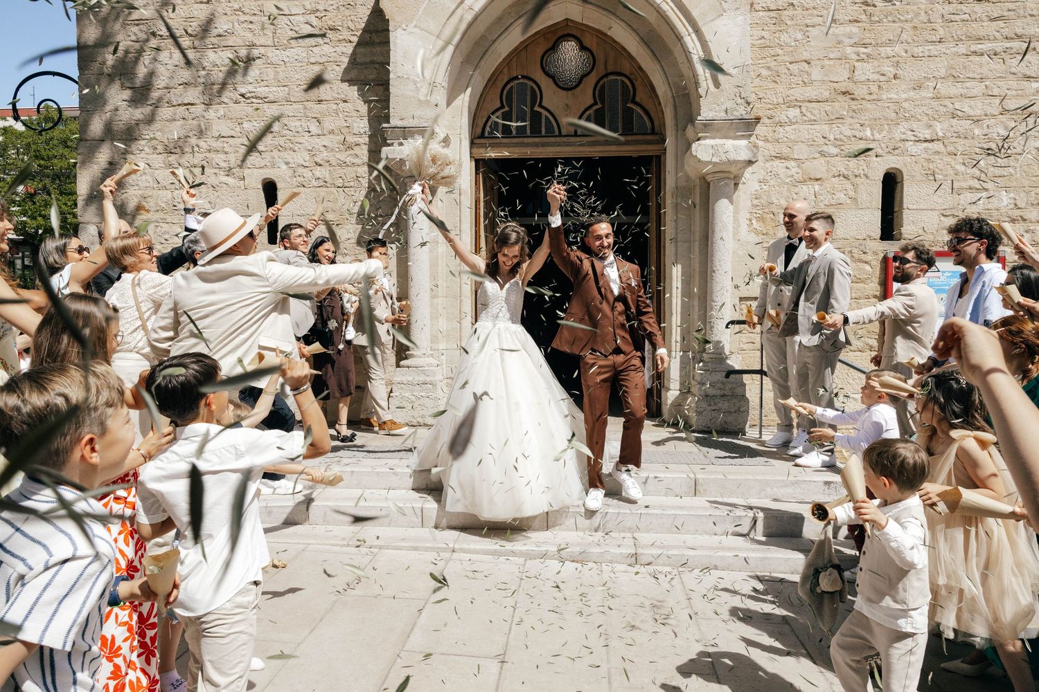 Newlywed couple exits stone church amid cheering guests throwing confetti on a sunny wedding day.