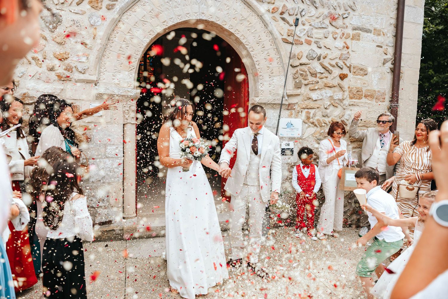 Bride and groom exit a stone church entrance as guests shower them with colorful confetti petals.