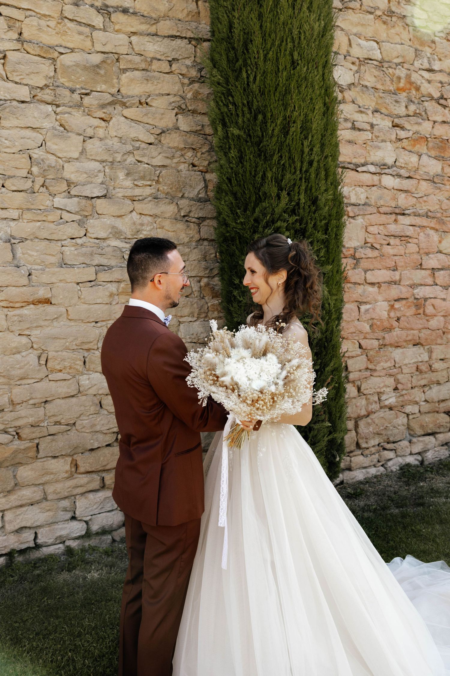 Bride and groom share a joyful moment before a stone wall, bride holding a beautiful dried floral bouquet.