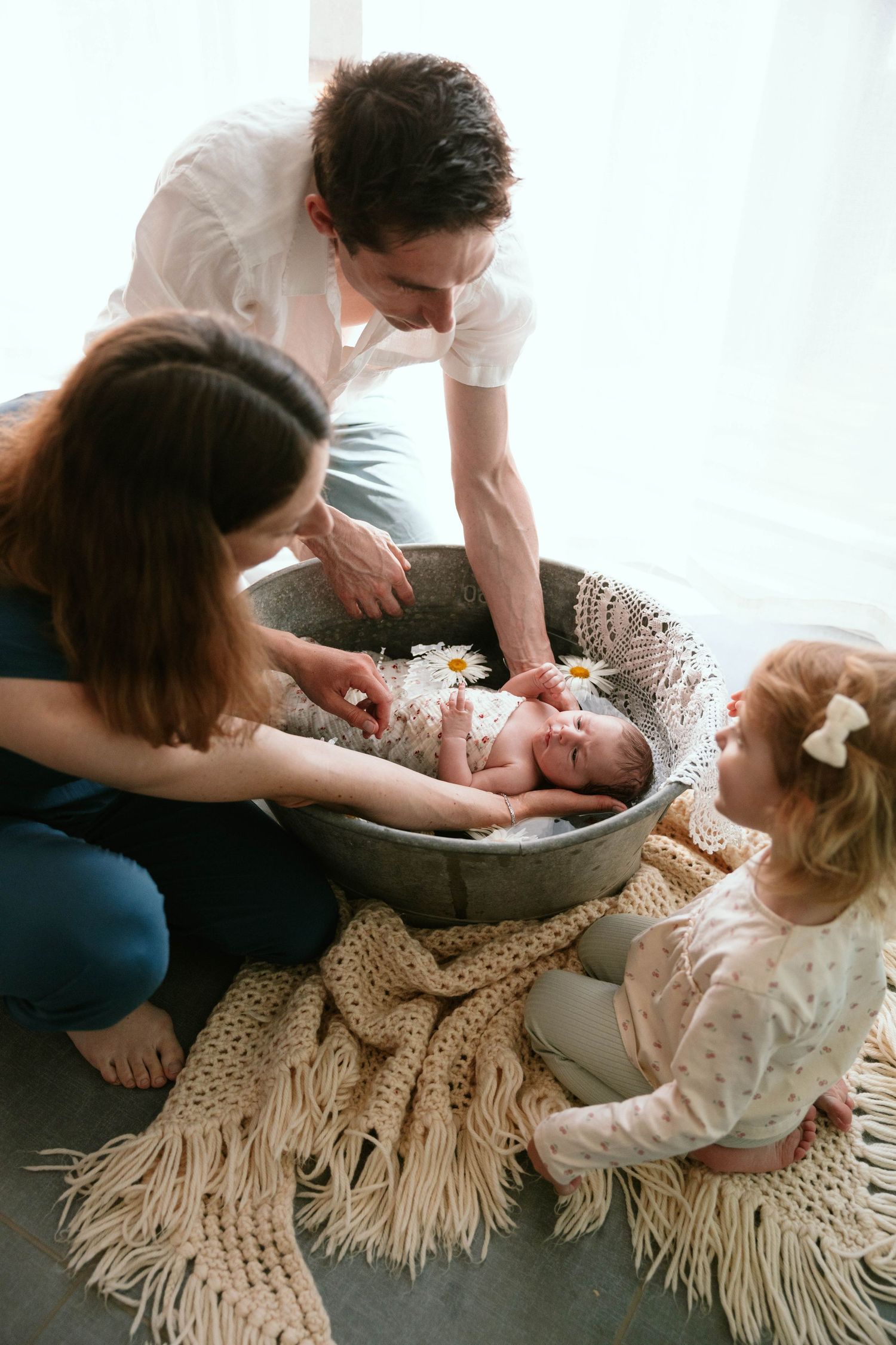 Parents and toddler daughter gather around newborn baby resting in a decorative bowl on the floor.