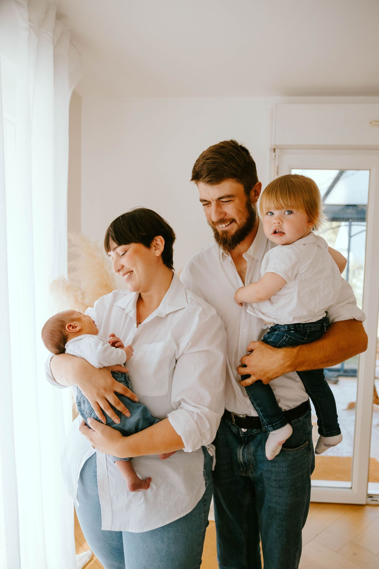 Happy family of four with newborn baby and toddler posing together in bright white home interior.