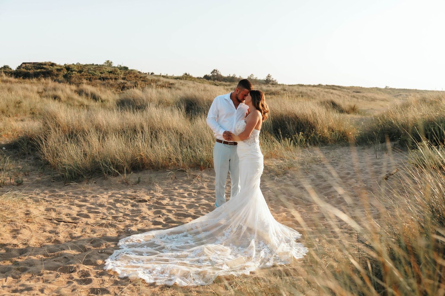 Bride in lace mermaid gown with long train kisses groom in white shirt on sandy beach dunes at sunset.