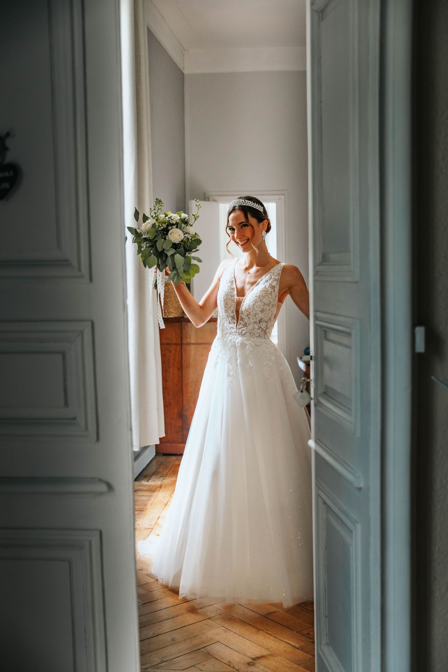 Smiling bride in white lace tulle gown holding green bouquet, standing in elegant doorway with herringbone floors.