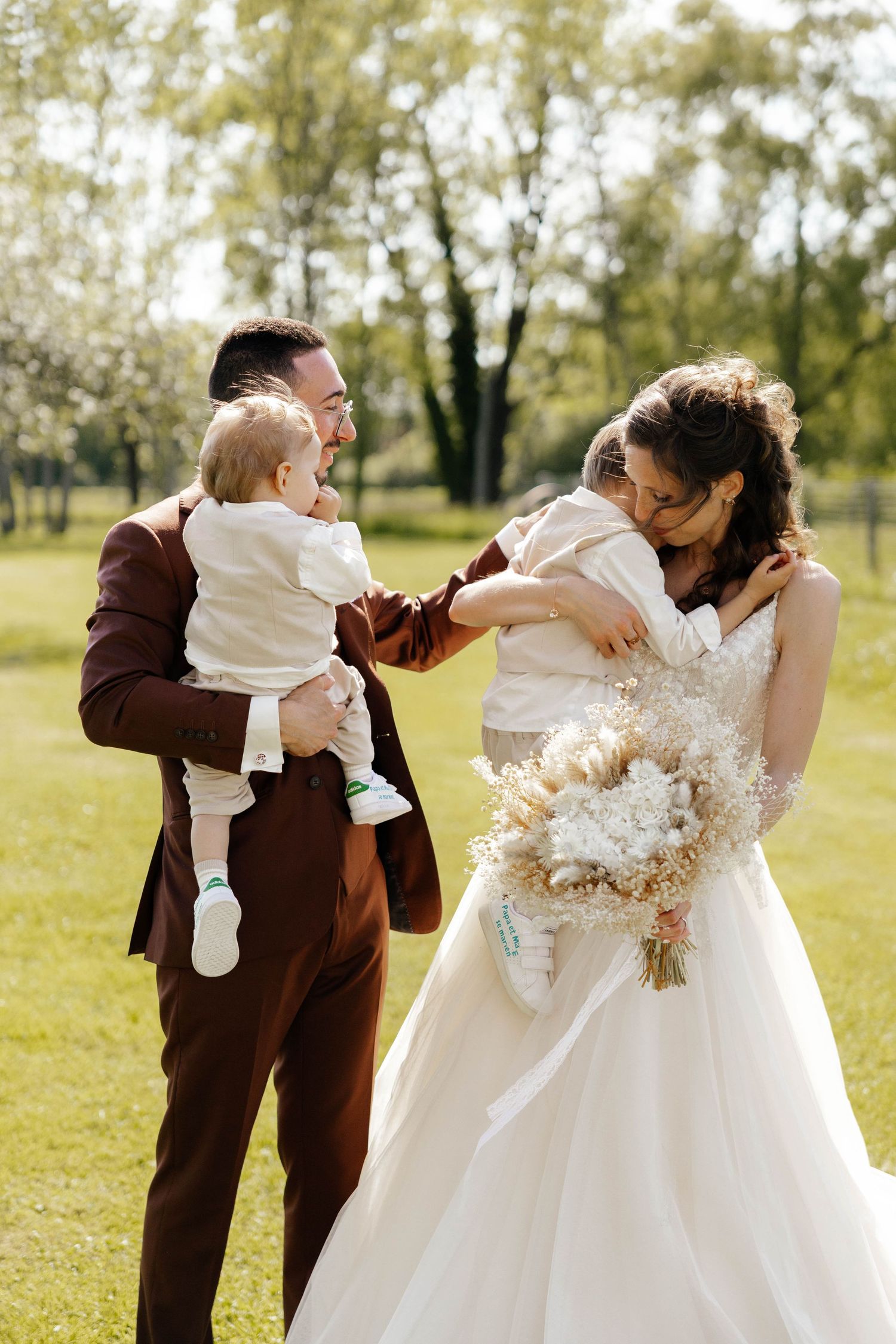 Happy bride and groom hold their two young children outdoors in a sunny green park on their wedding day.