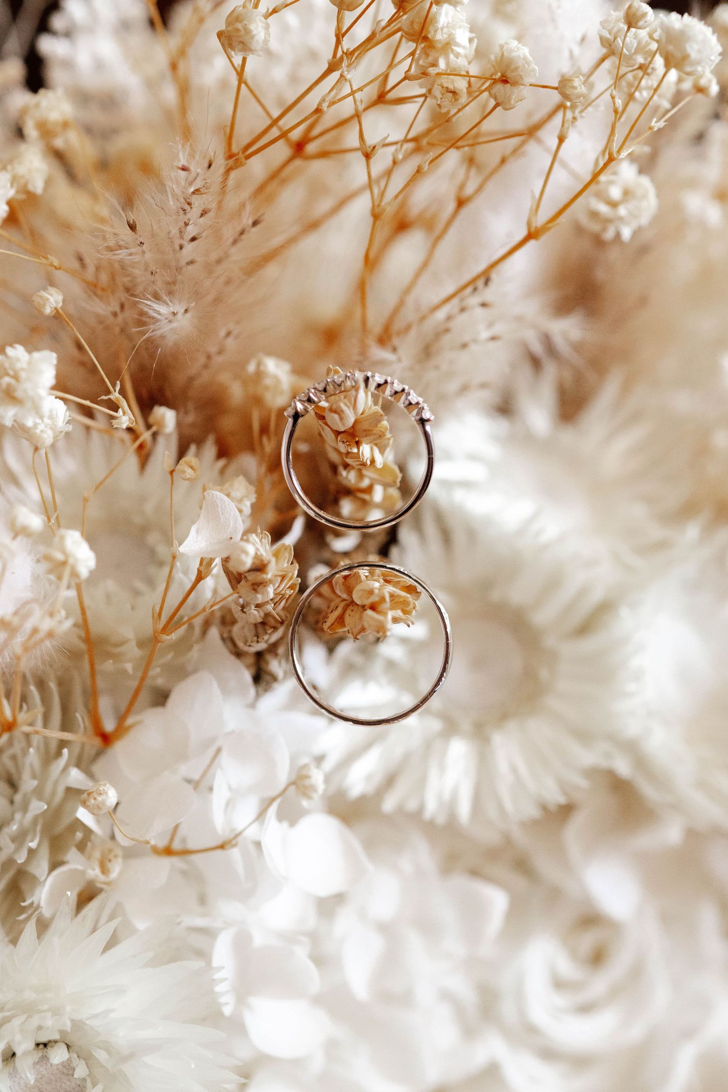Two delicate diamond wedding rings nestled among dried white and beige flowers in a bridal bouquet.