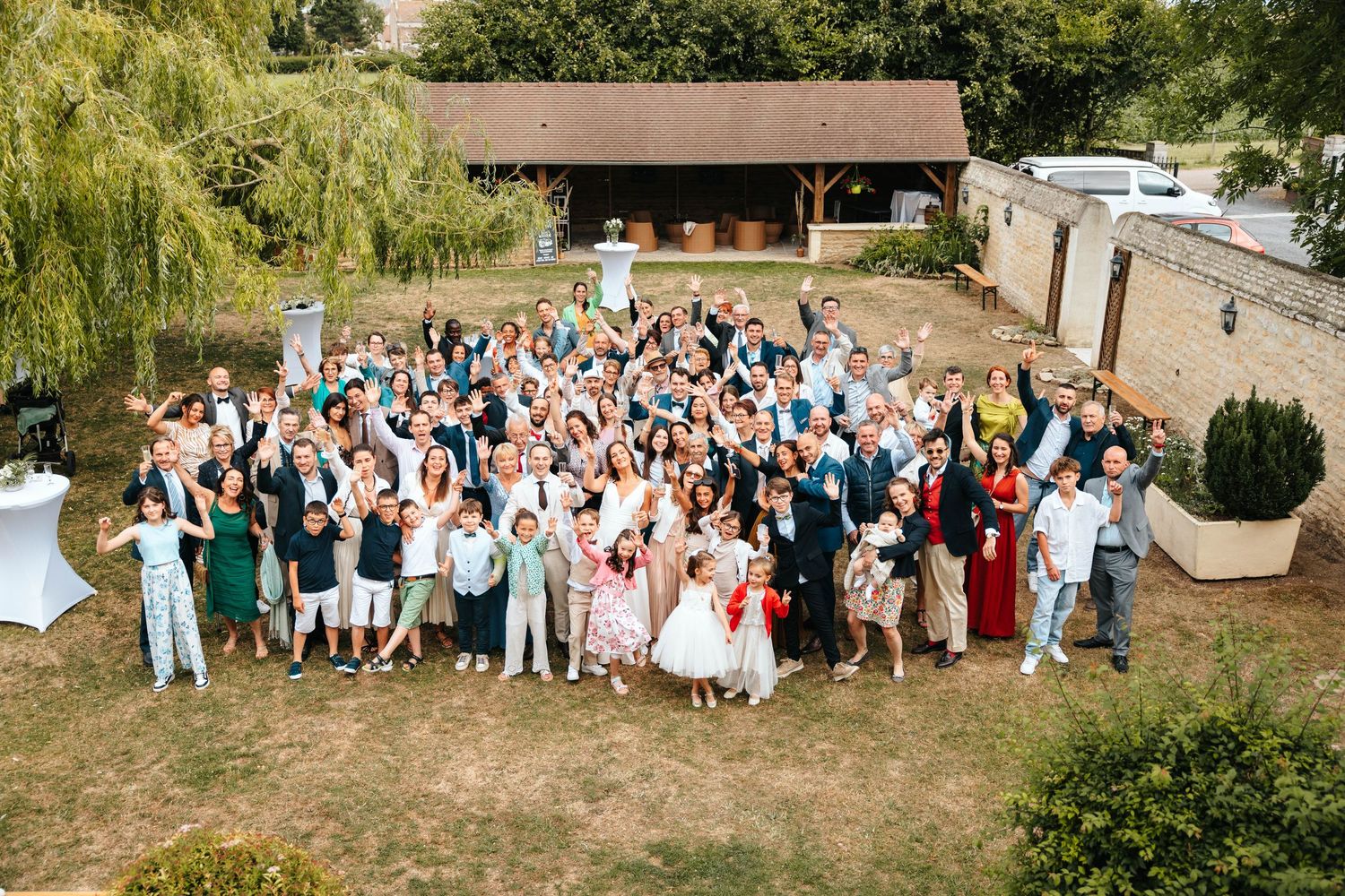 Large wedding party group photo taken from above showing all guests celebrating outdoors on a sunny day.