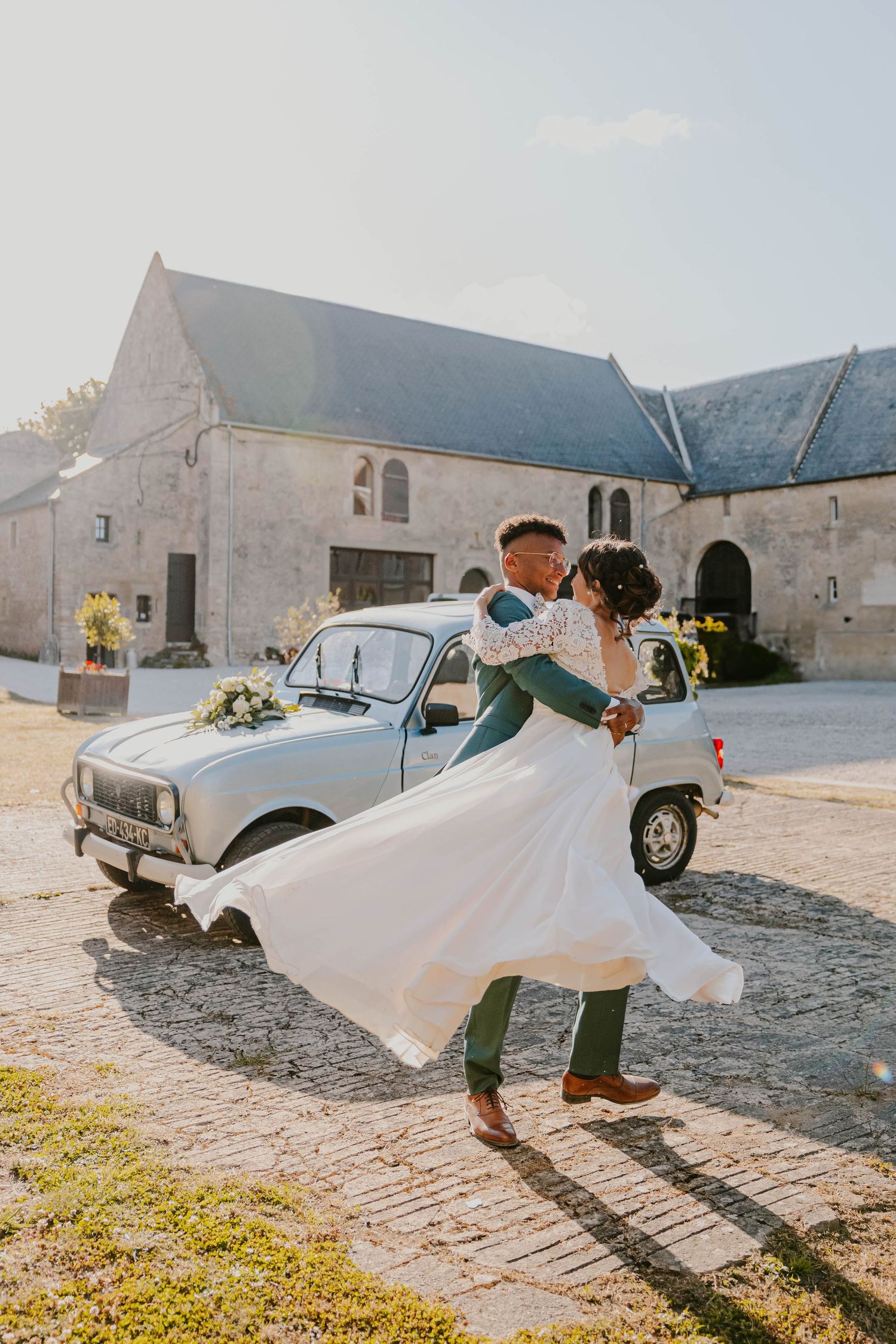 Newlywed couple sharing a romantic dip kiss in front of a vintage Renault 4 at a rustic French countryside wedding venue.