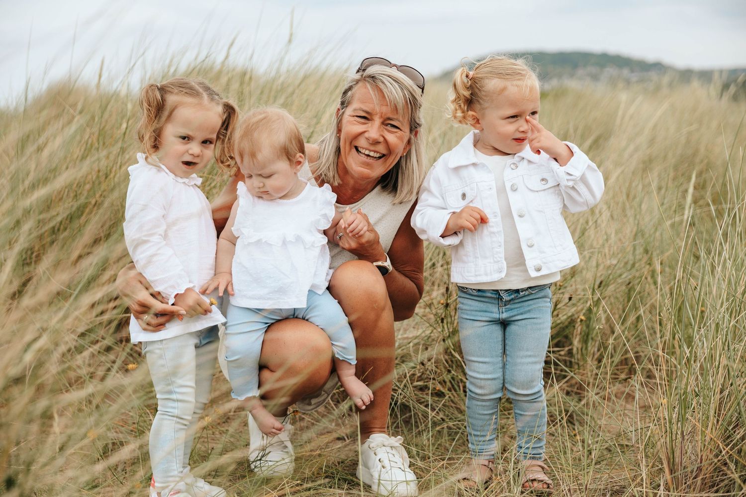 Woman laughing with three young children in white outfits among beach grass dunes.