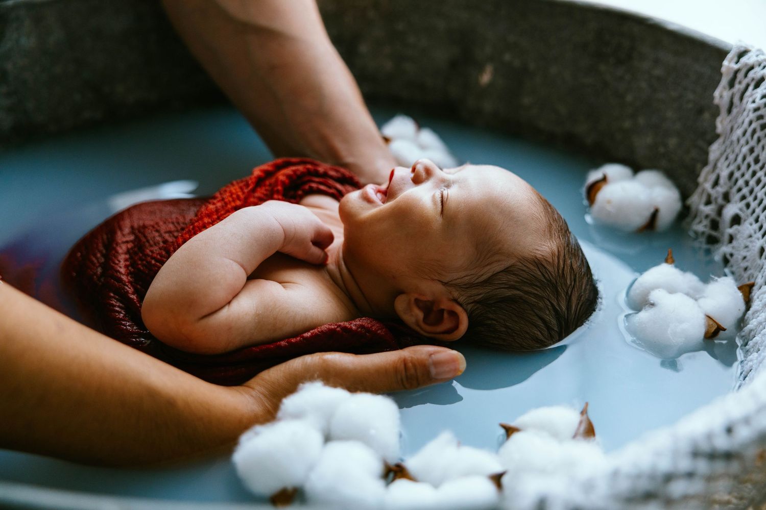 Newborn baby wrapped in red cloth floating in water surrounded by cotton flowers.