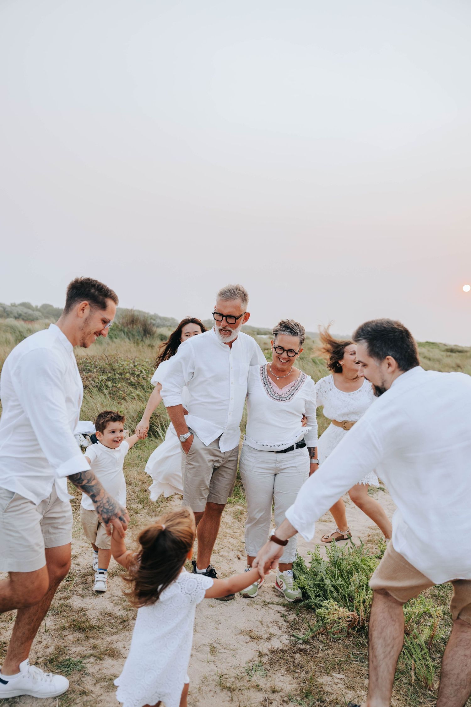 Multigenerational family in white clothing playing together on a sandy coastal path at sunset.