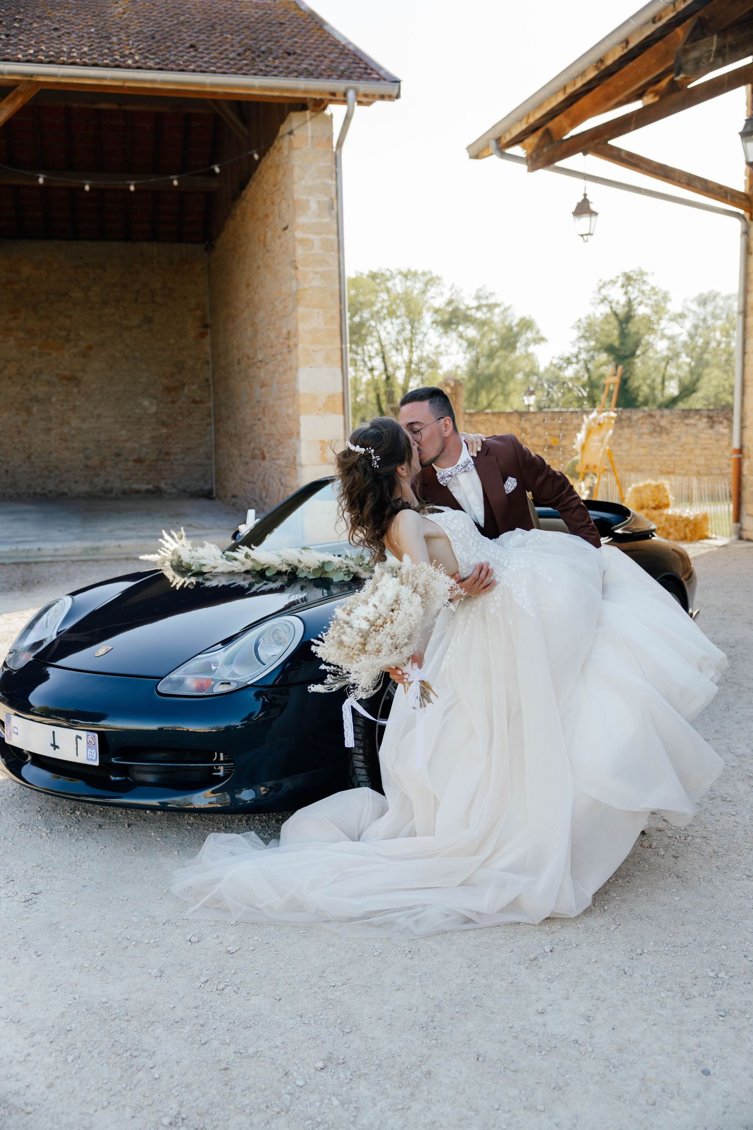 Bride and groom kissing beside decorated black Porsche sports car at rustic wedding venue.