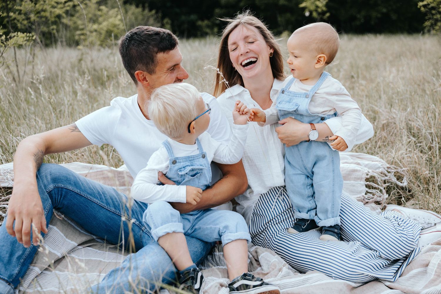 Happy family with two toddlers laughing on a blanket in a sunny meadow, wearing white shirts and denim.