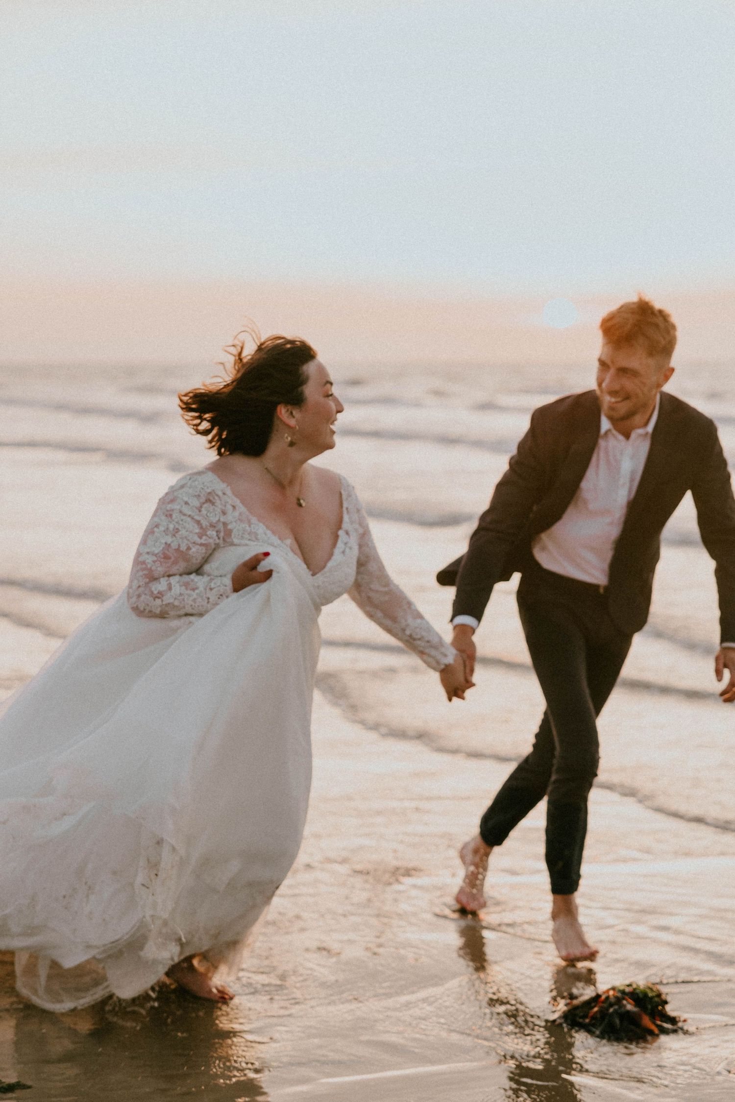 Bride and groom laughing and running hand-in-hand along a sandy beach at sunset in wedding attire.