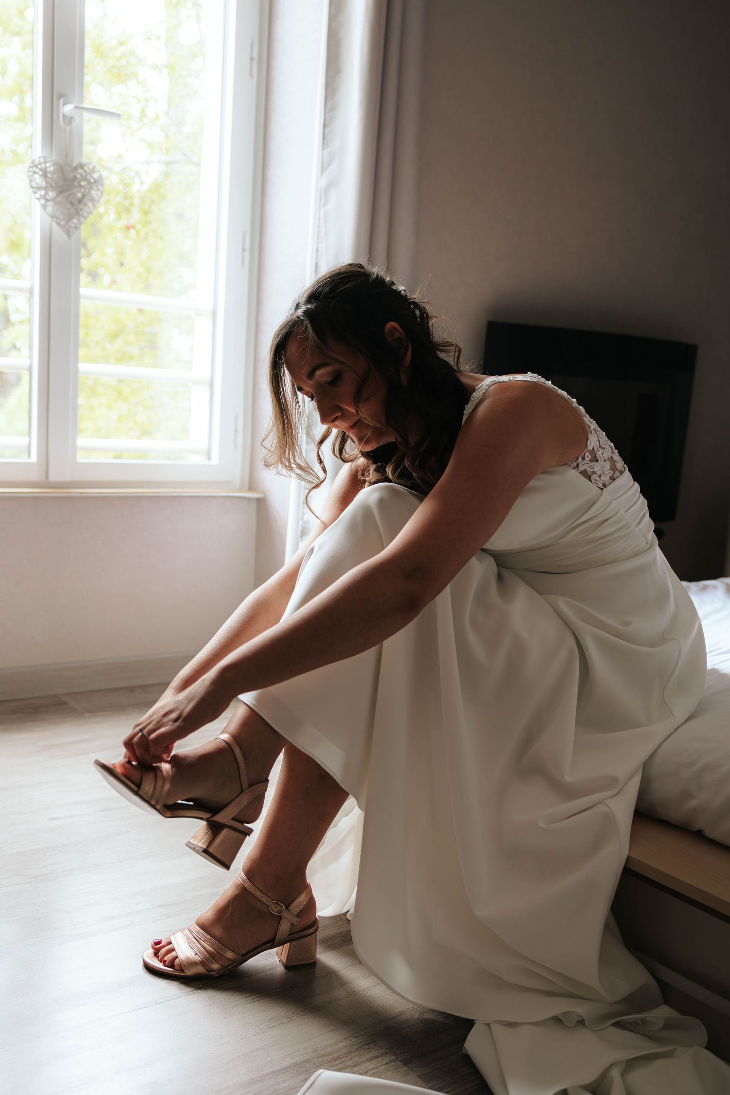 Bride in white lace wedding dress sitting by window putting on gold strappy heels before ceremony.