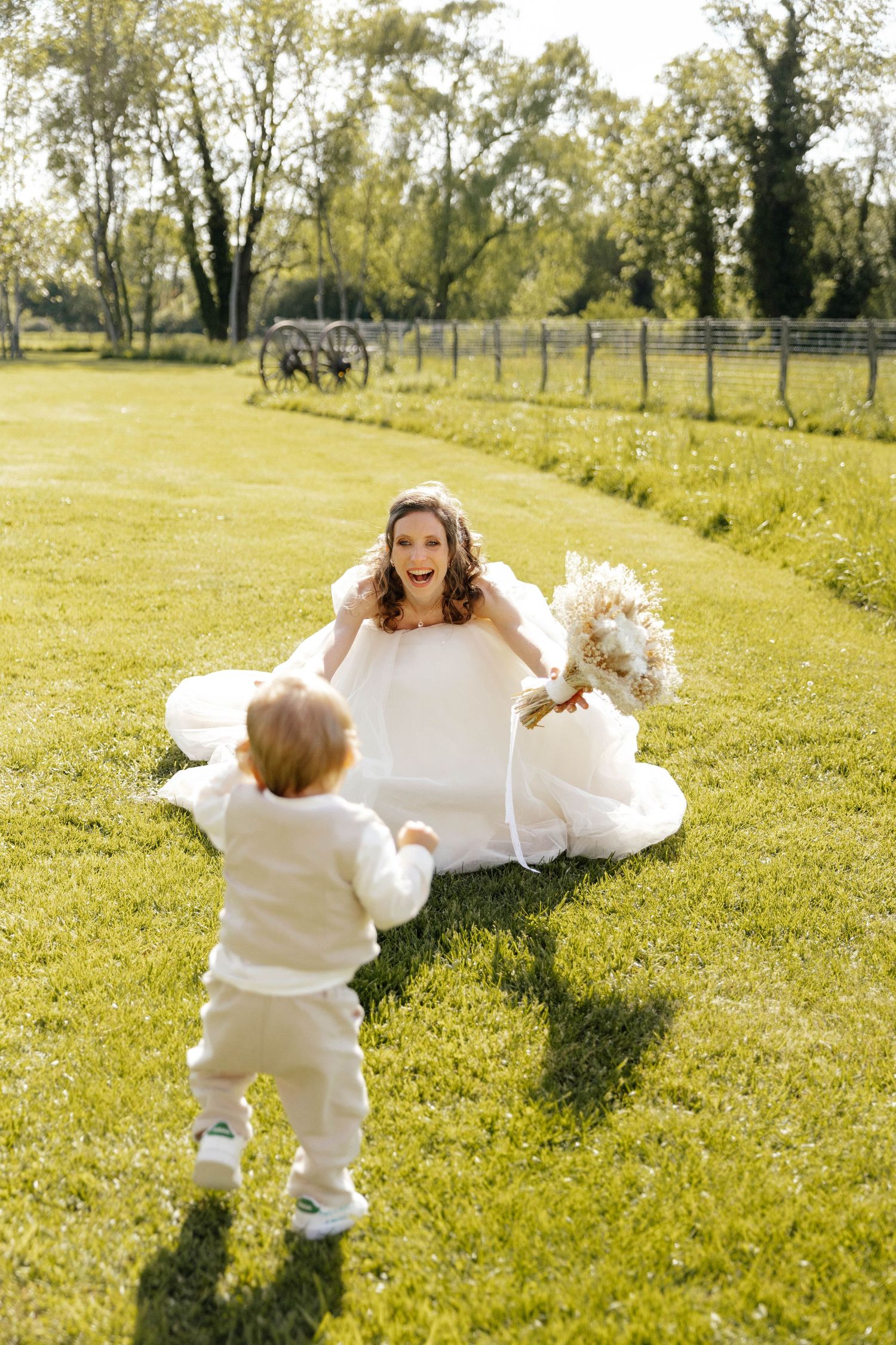 Happy bride in white gown sitting on grass welcoming toddler with bouquet outdoors.