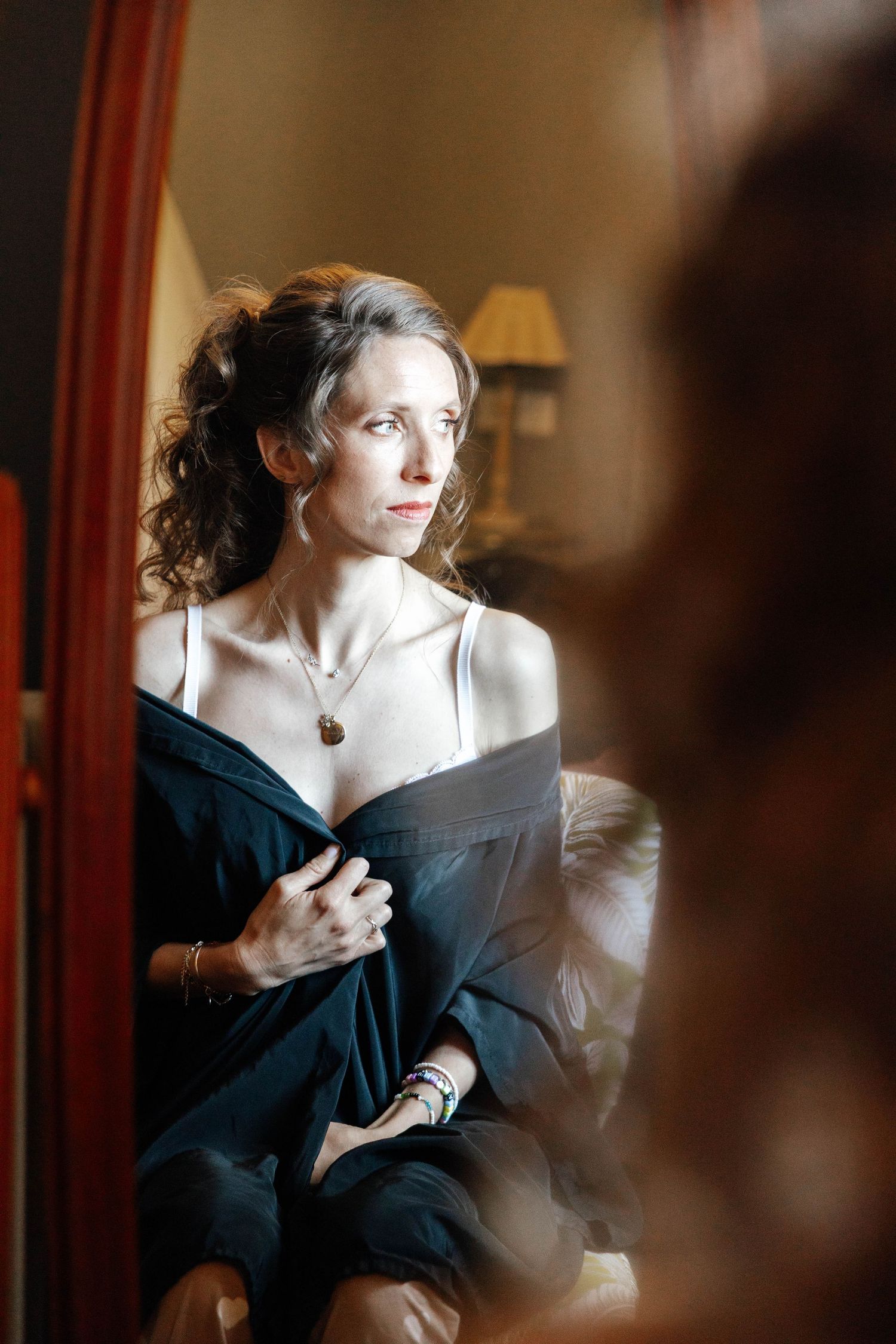 Elegant woman in dark robe and necklace sitting thoughtfully by a mirror in a warmly lit room.