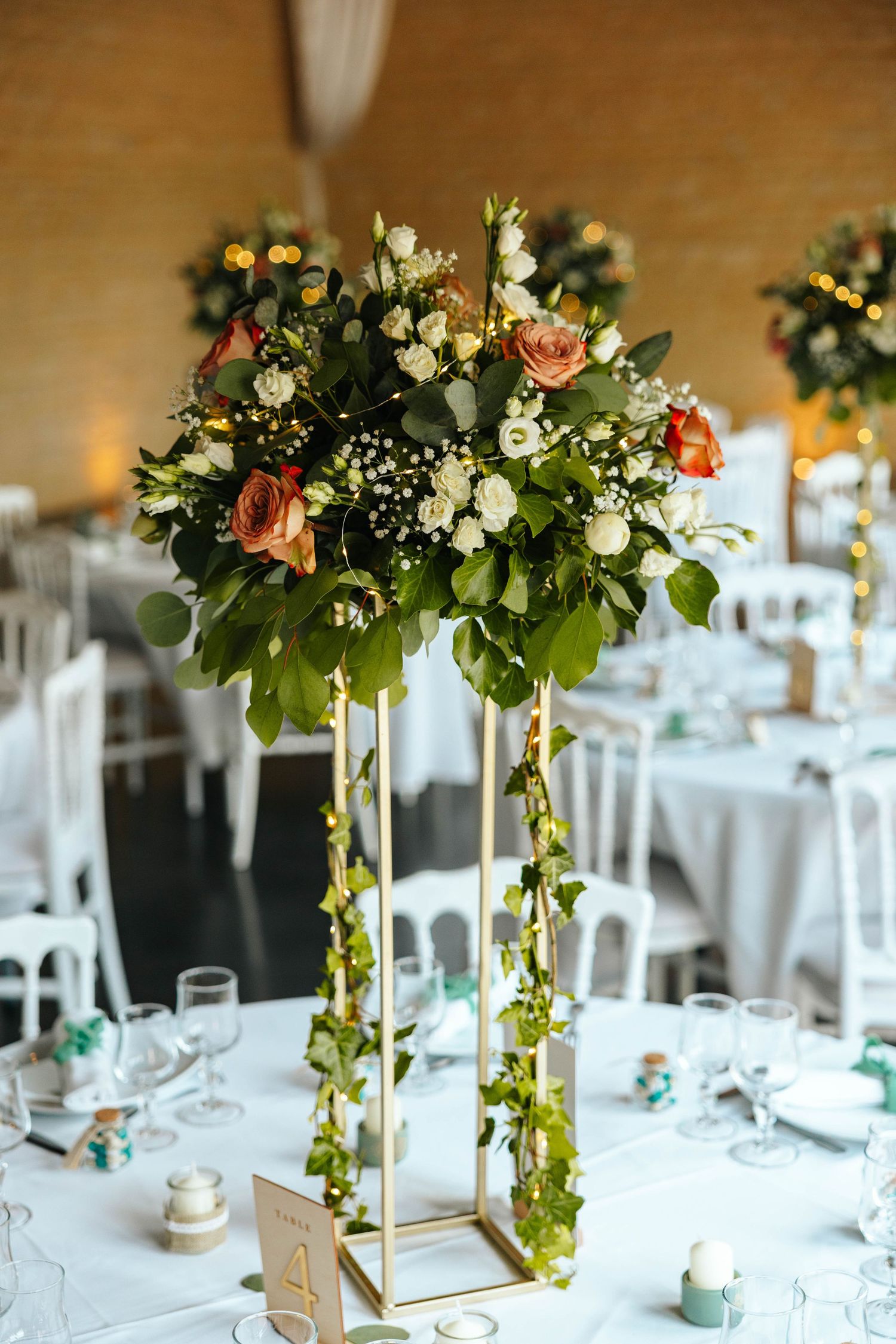 Tall gold geometric vase with lush floral centerpiece of orange roses, white blooms, and trailing greenery on wedding table.