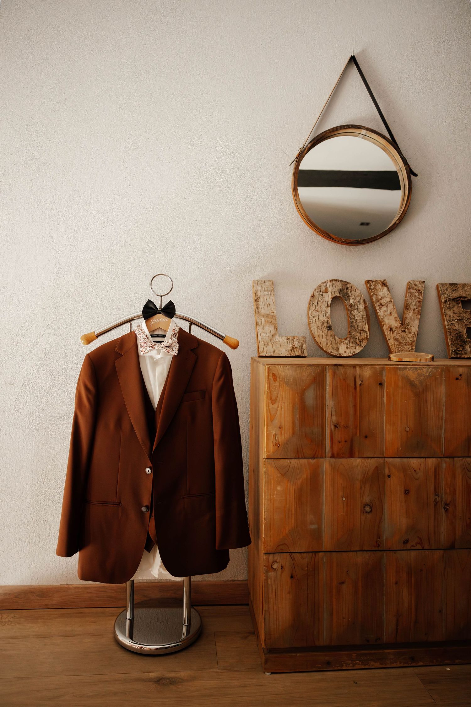 Brown suit jacket on hanger beside wooden dresser with LOVE letters and round mirror on white wall.
