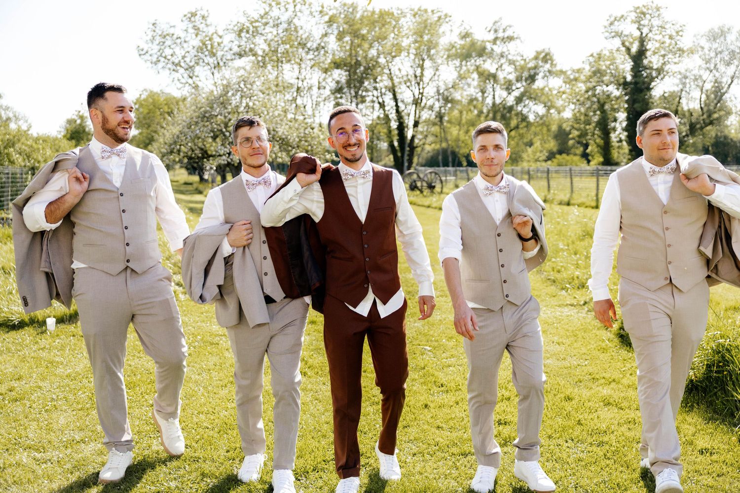 Groom in brown suit poses with four groomsmen in beige vests on sunny lawn.