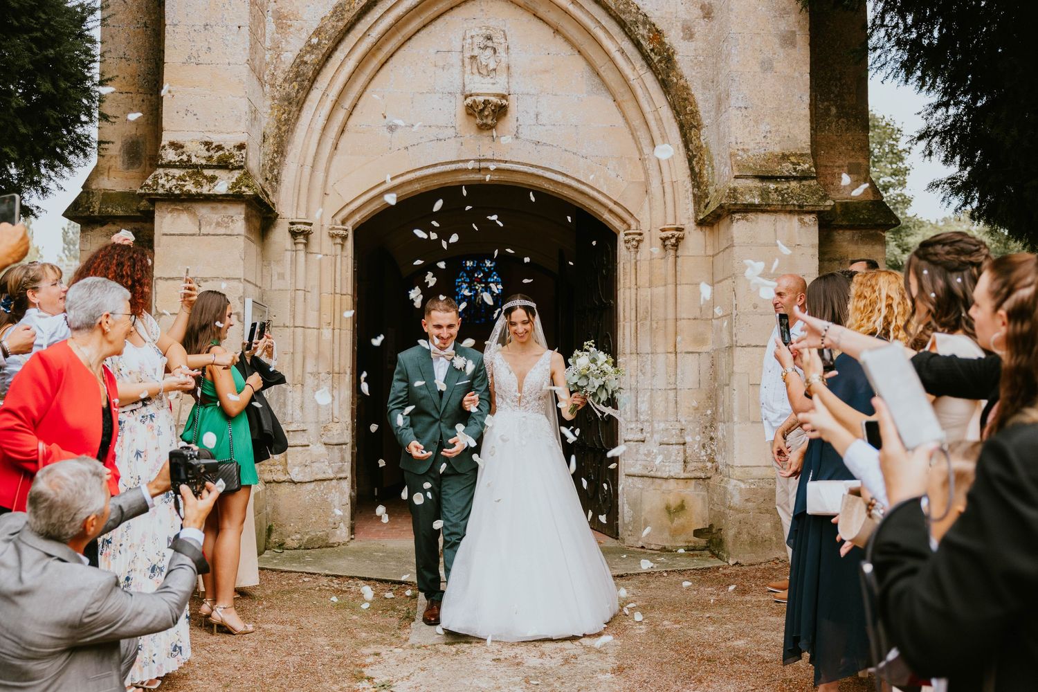 Bride and groom exit stone church as guests throw confetti at outdoor wedding ceremony.