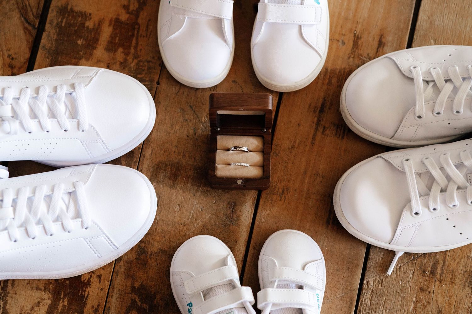 White sneakers and a wooden ring box with wedding rings arranged on a rustic wooden floor.