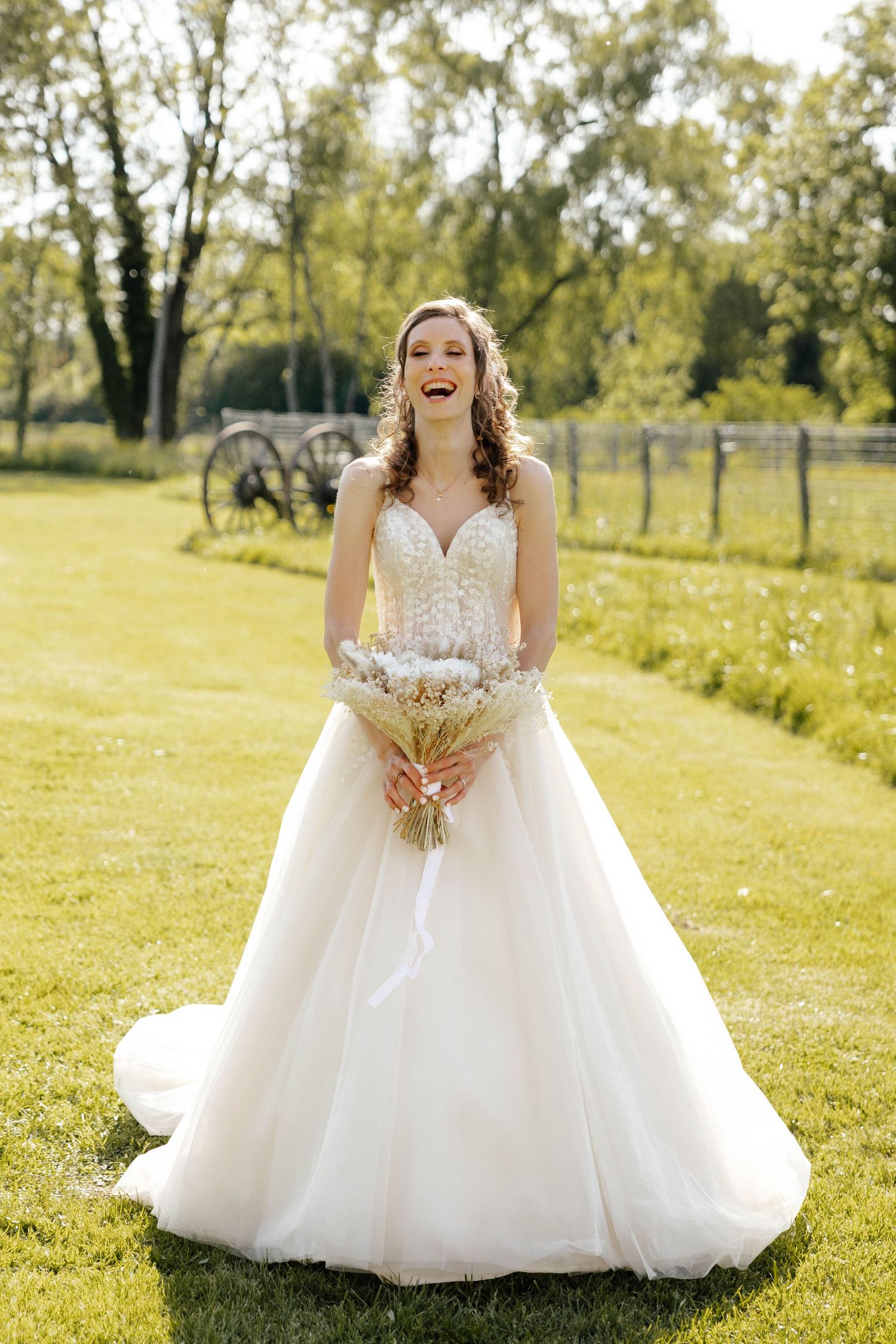 Joyful bride in elegant white ballgown laughing while holding bouquet on green lawn with trees in background.