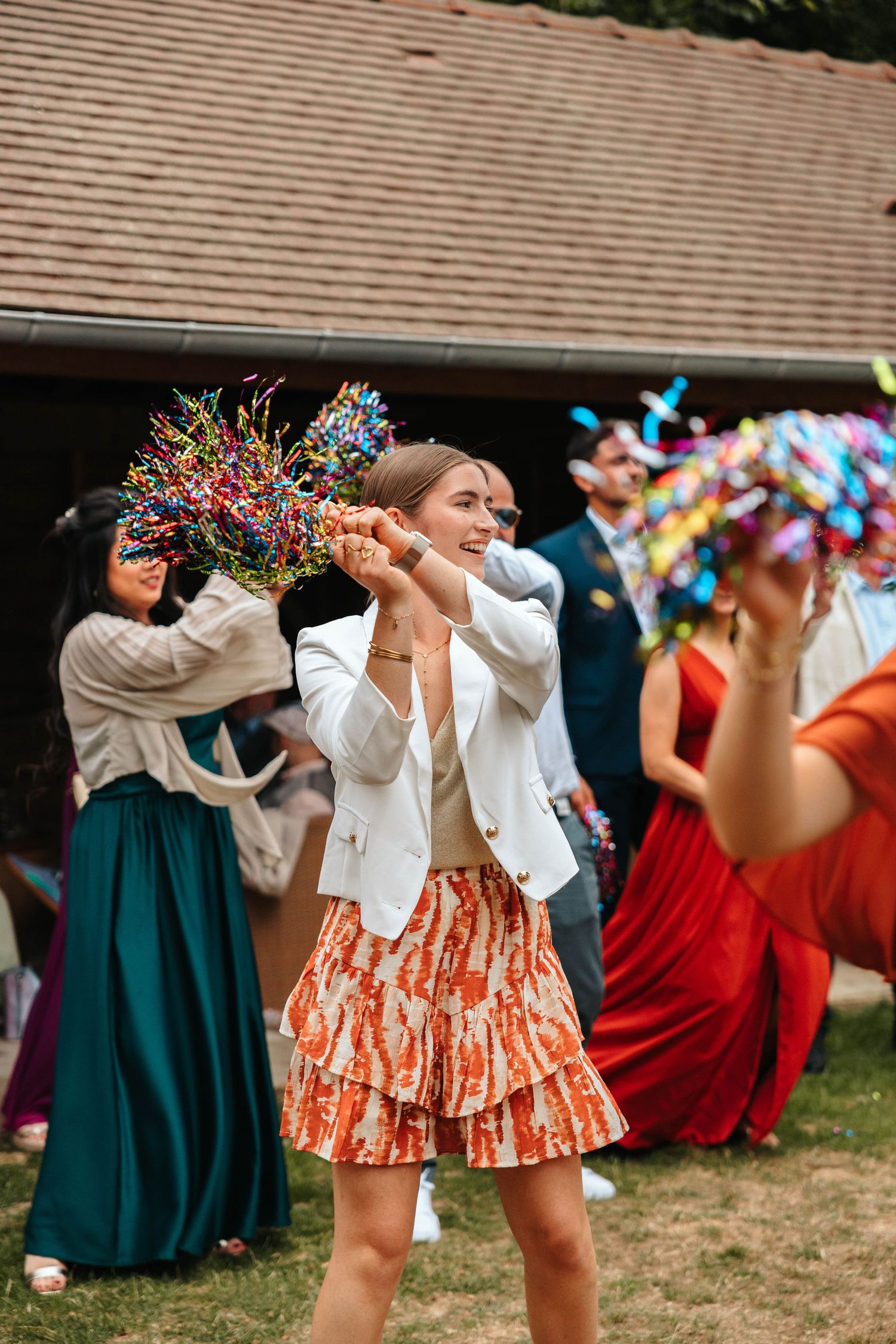 Smiling wedding guest in white blazer and floral skirt waves colorful confetti bouquet during outdoor celebration.