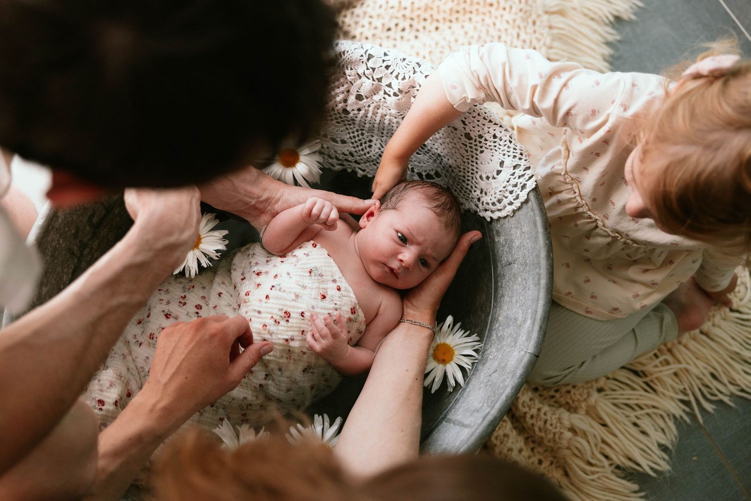 Overhead view of newborn baby in vintage bowl with daisies as family hands reach in lovingly.