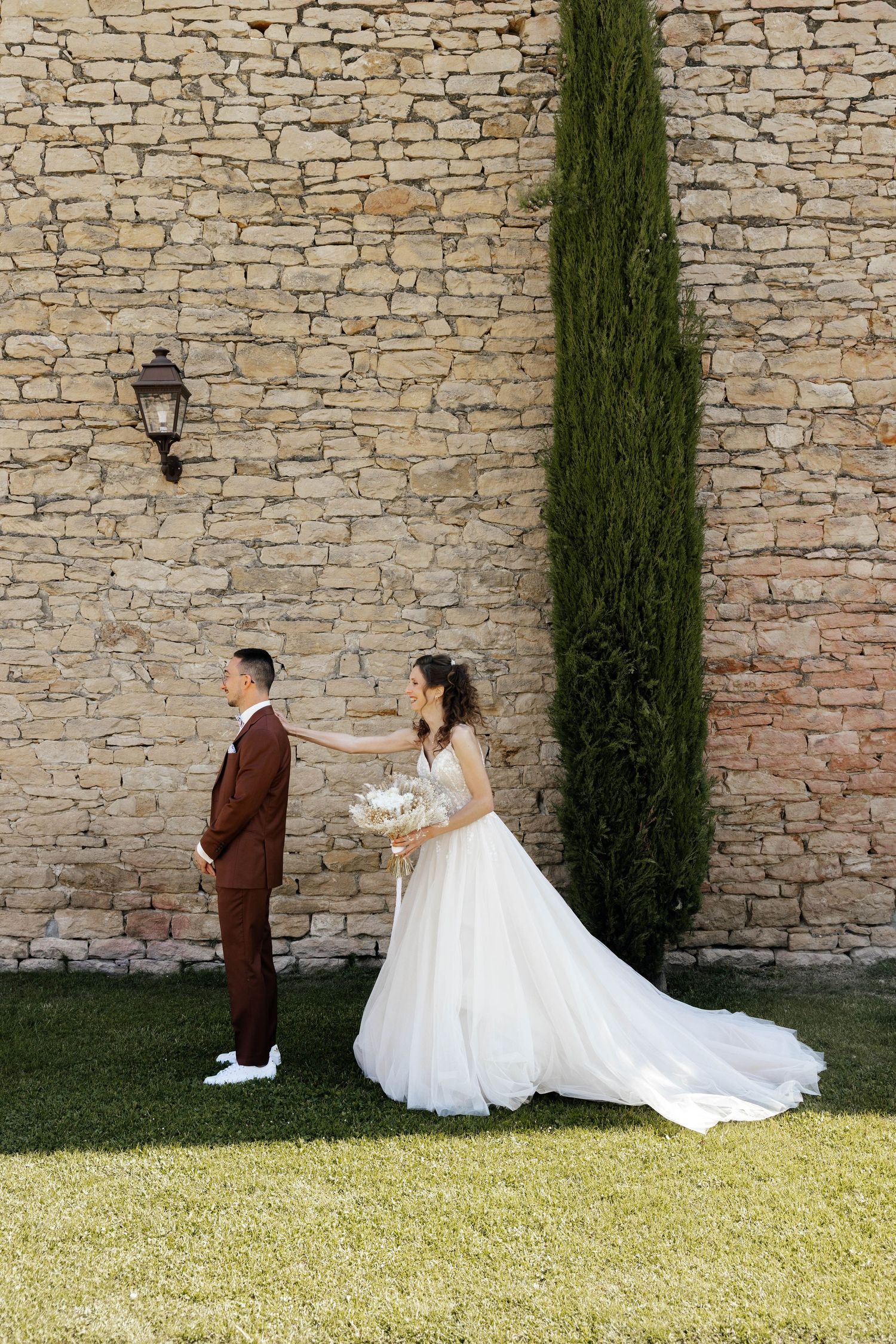 Bride in flowing white dress taps groom on shoulder during first look moment beside stone wall and cypress tree.