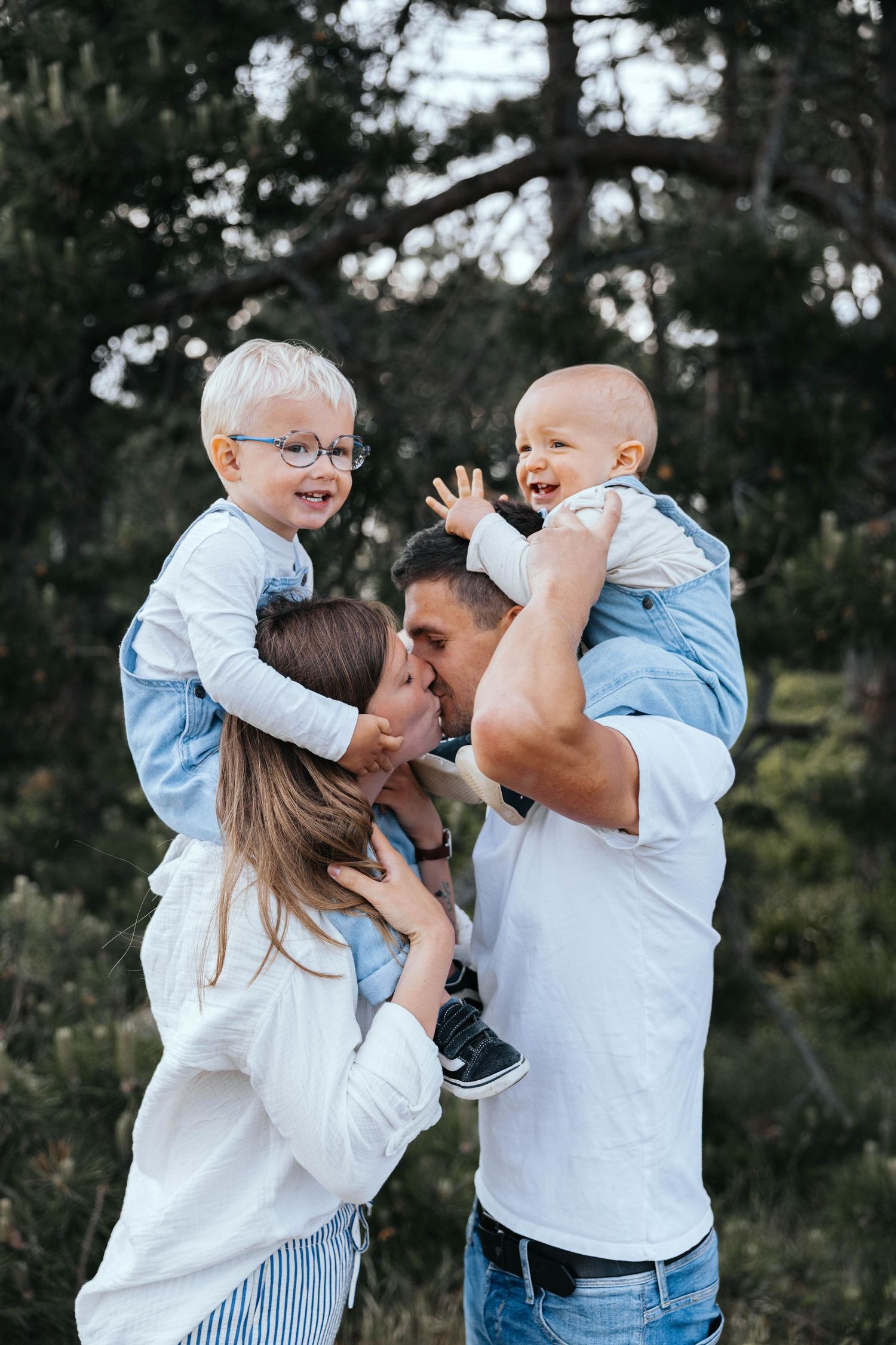 Happy family with two young boys outdoors, parents kissing while holding their children in a lush green setting.