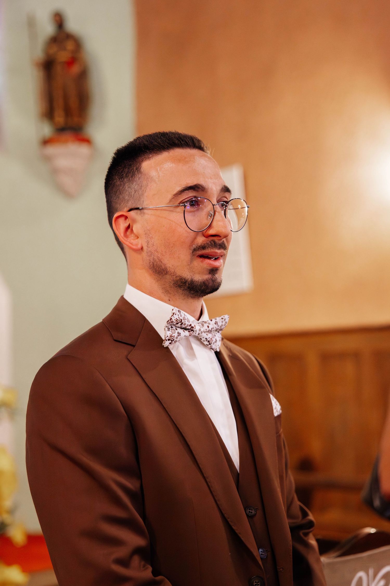 Groom in brown suit and floral bow tie standing attentively inside a church.