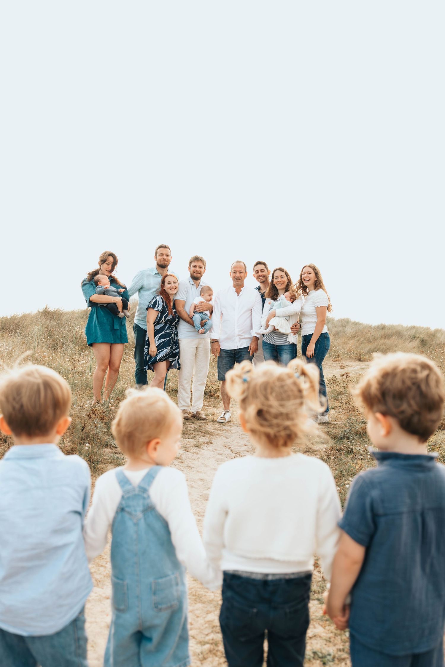 Young children in overalls walk toward a large multigenerational family gathered on a sandy dune path.