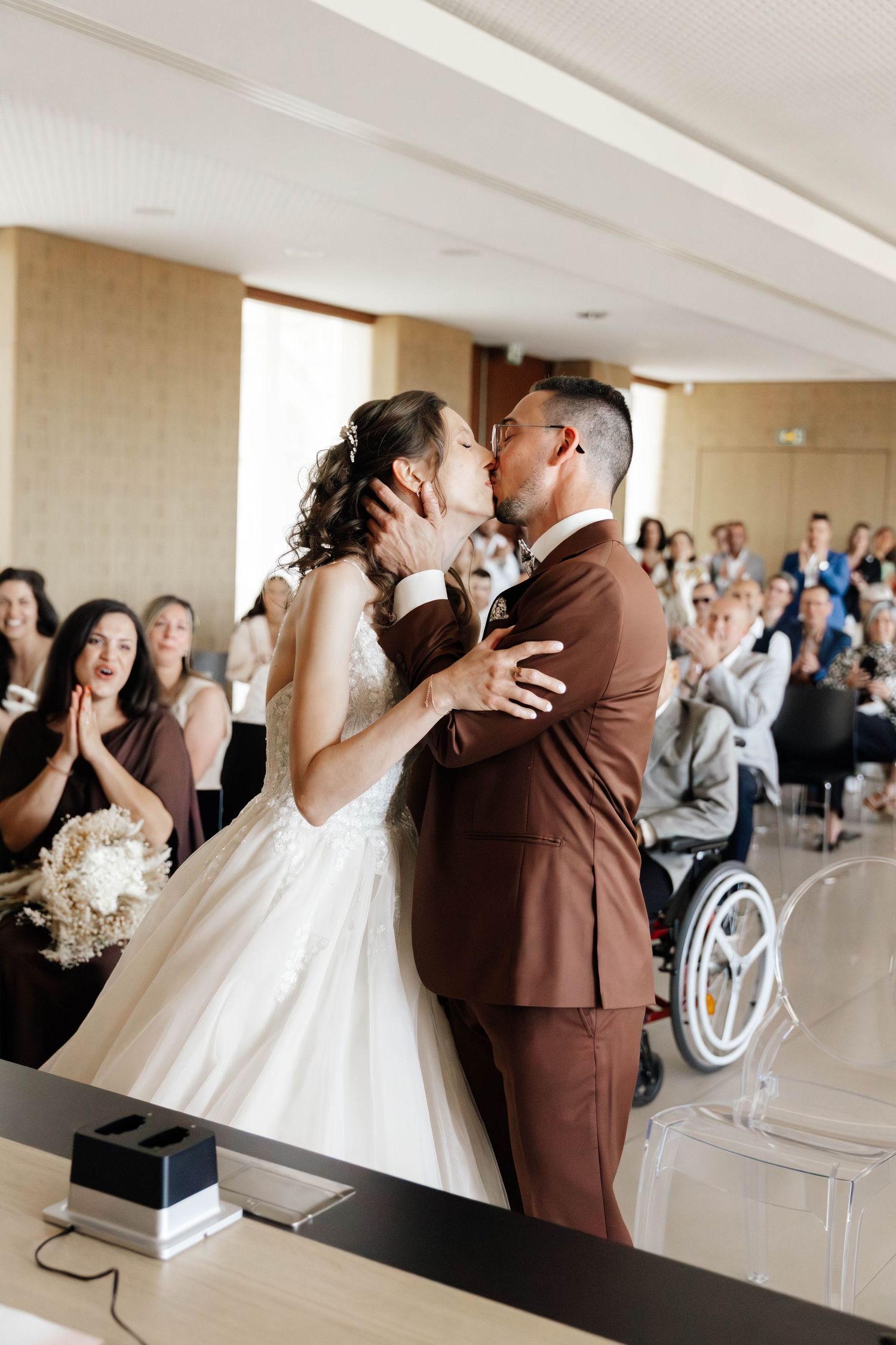Bride and groom share their first kiss at wedding ceremony while guests applaud in bright modern venue.