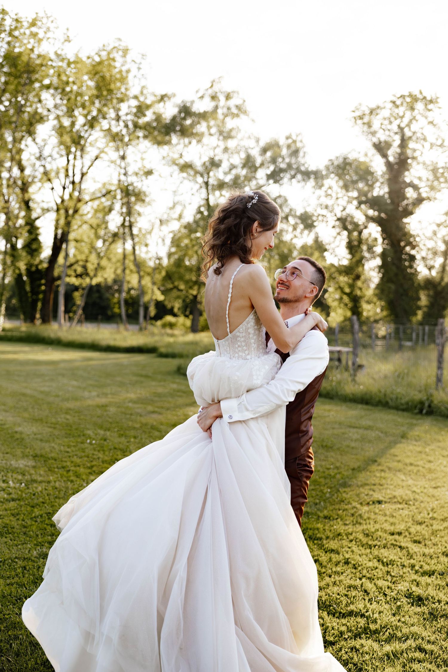 Joyful groom lifting his bride in a white gown outdoors in golden sunset light.