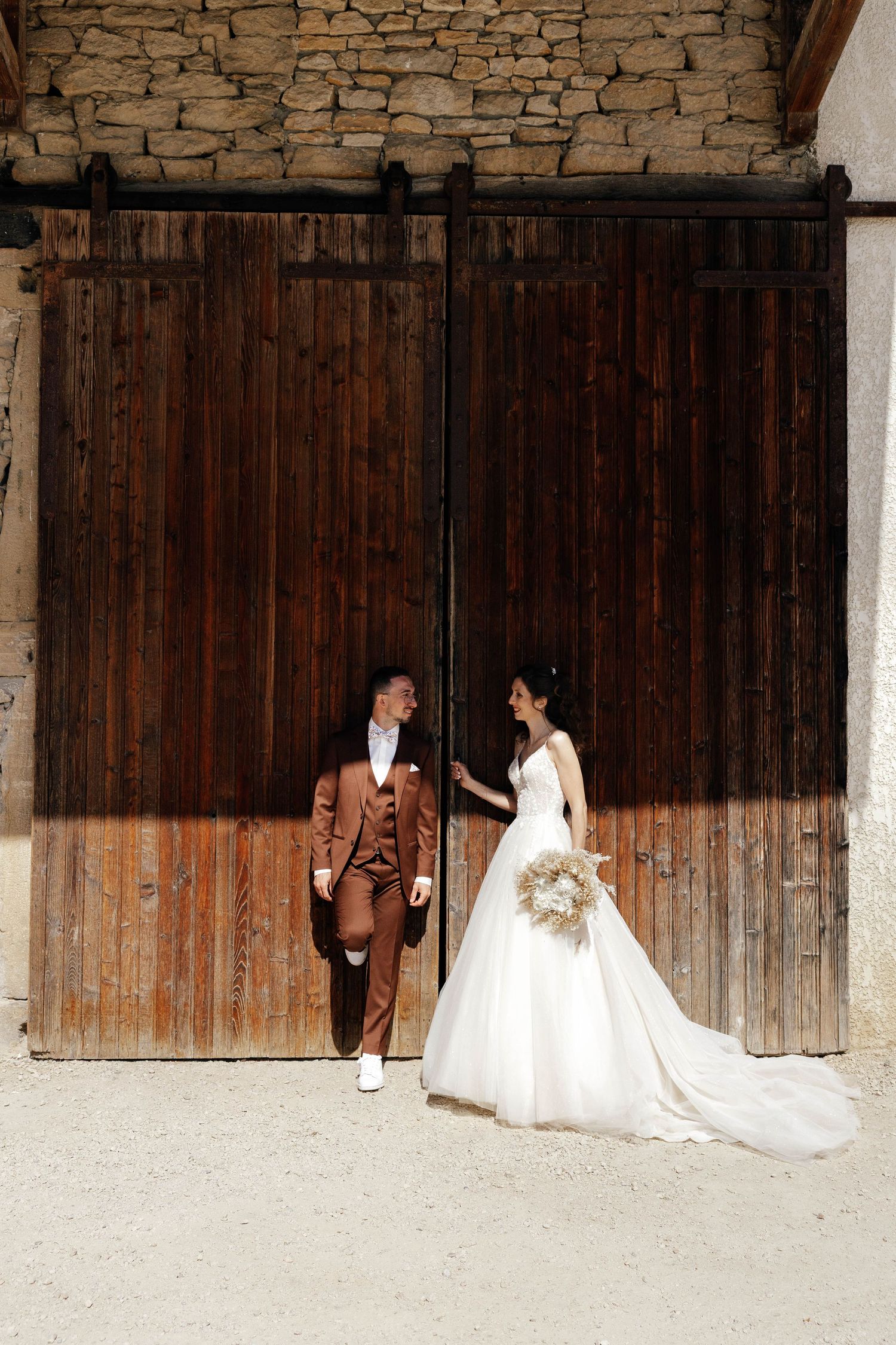 Wedding couple standing before rustic wooden barn doors, bride in white gown holding bouquet.