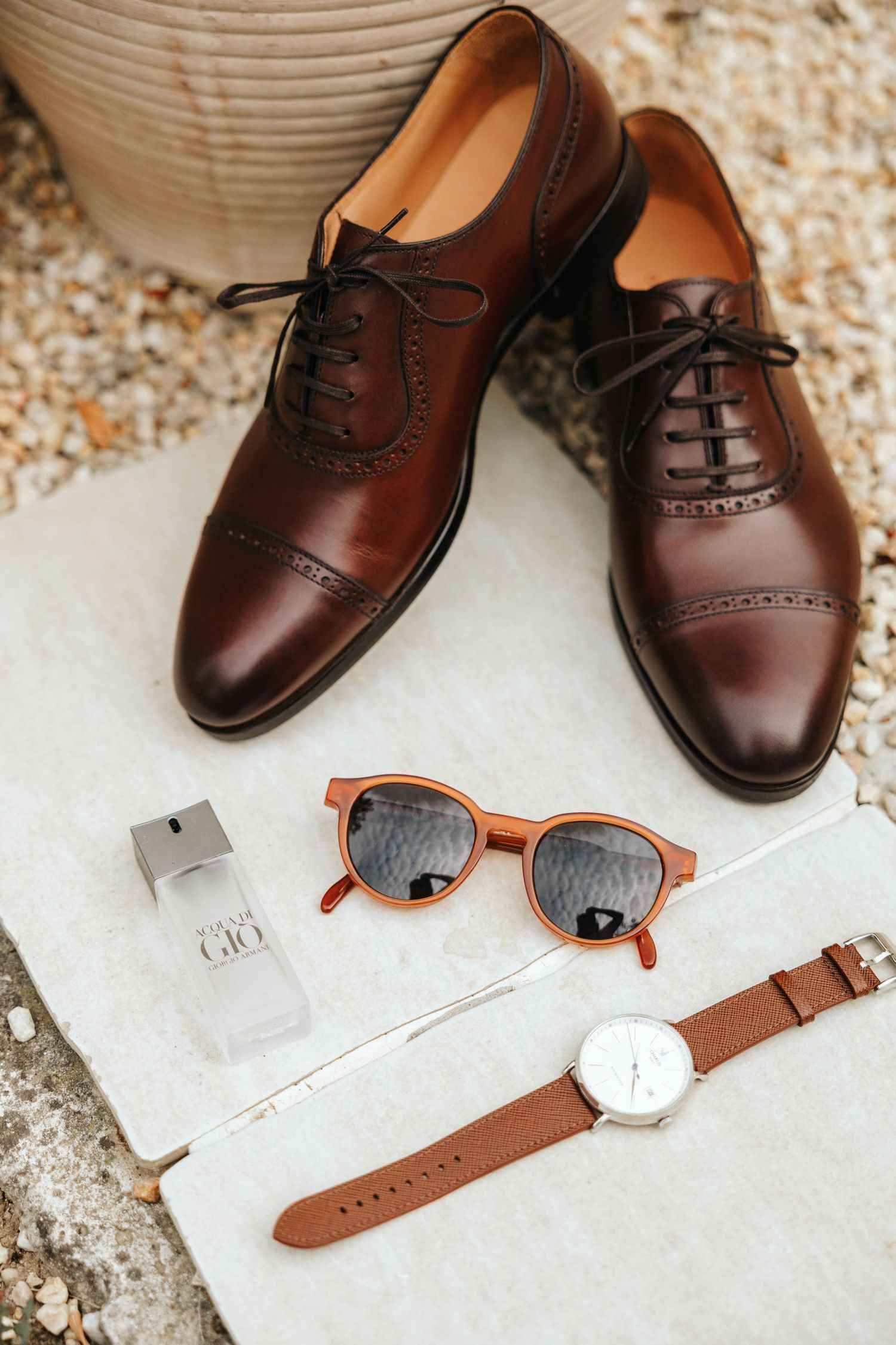Groom accessories flat lay featuring brown Oxford shoes, sunglasses, watch, and cologne on white surface.