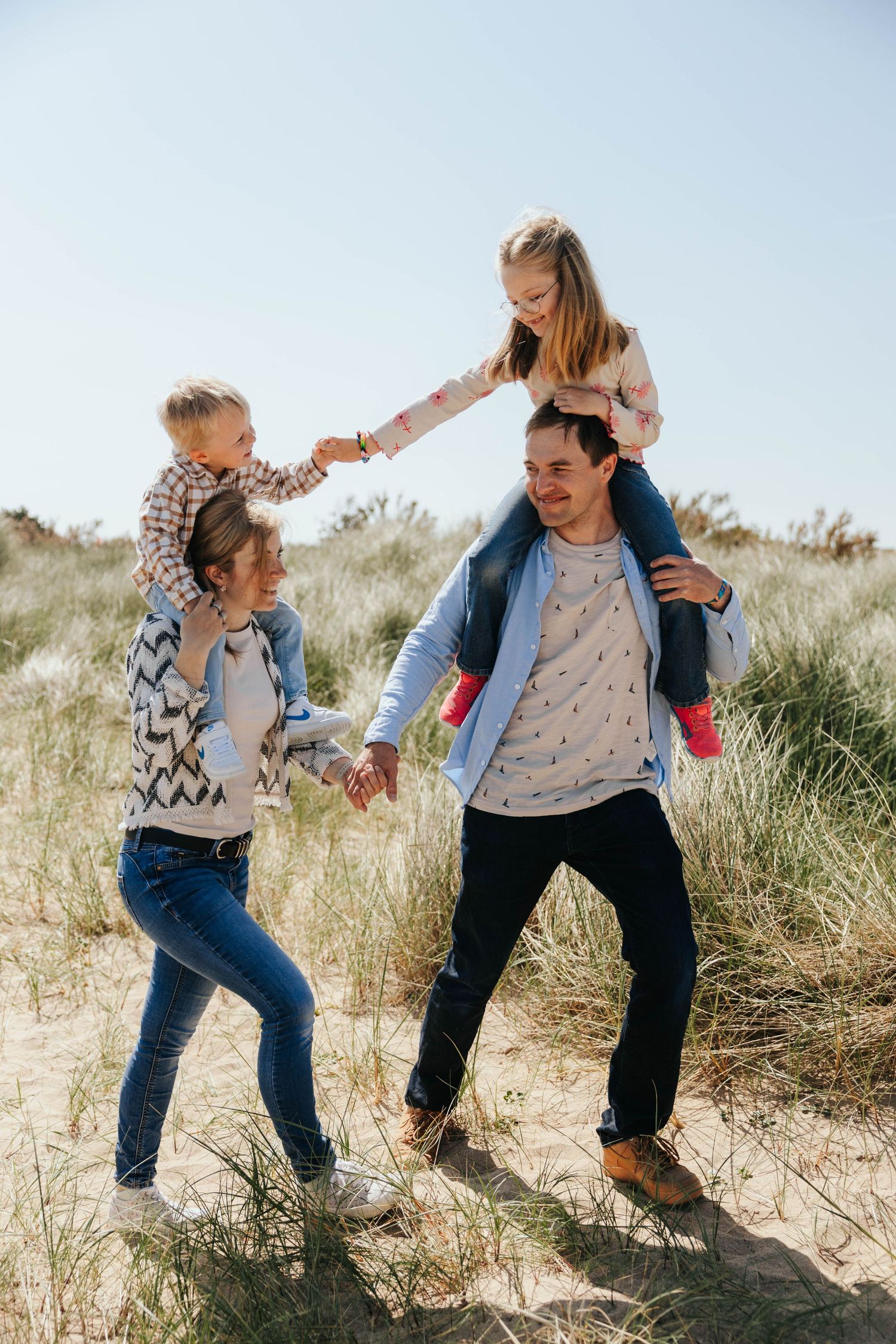 A family of four walks through sandy beach dunes, with children riding on parents shoulders on a sunny day.