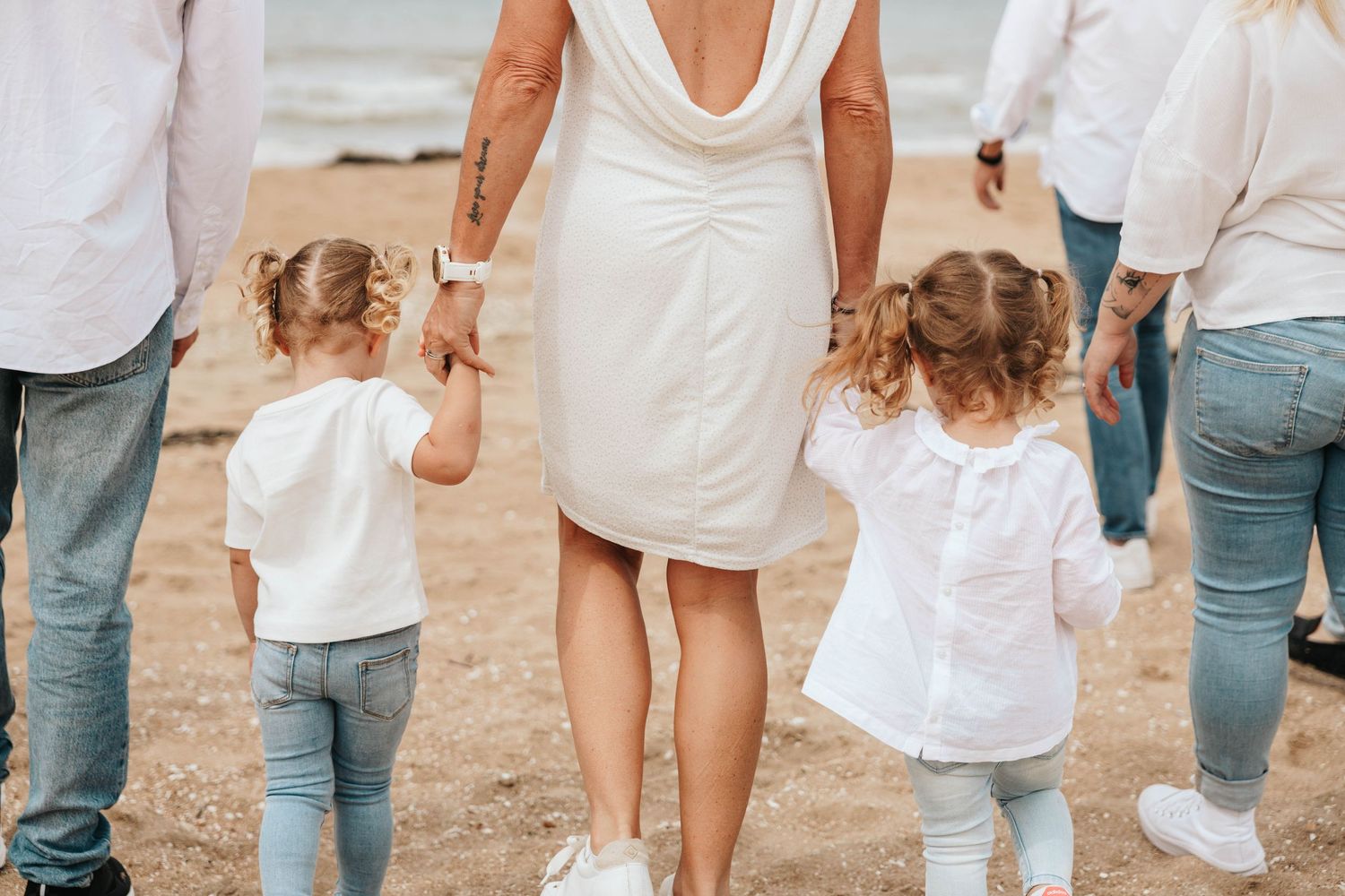 Family walking on beach holding hands, view from behind in white outfits.