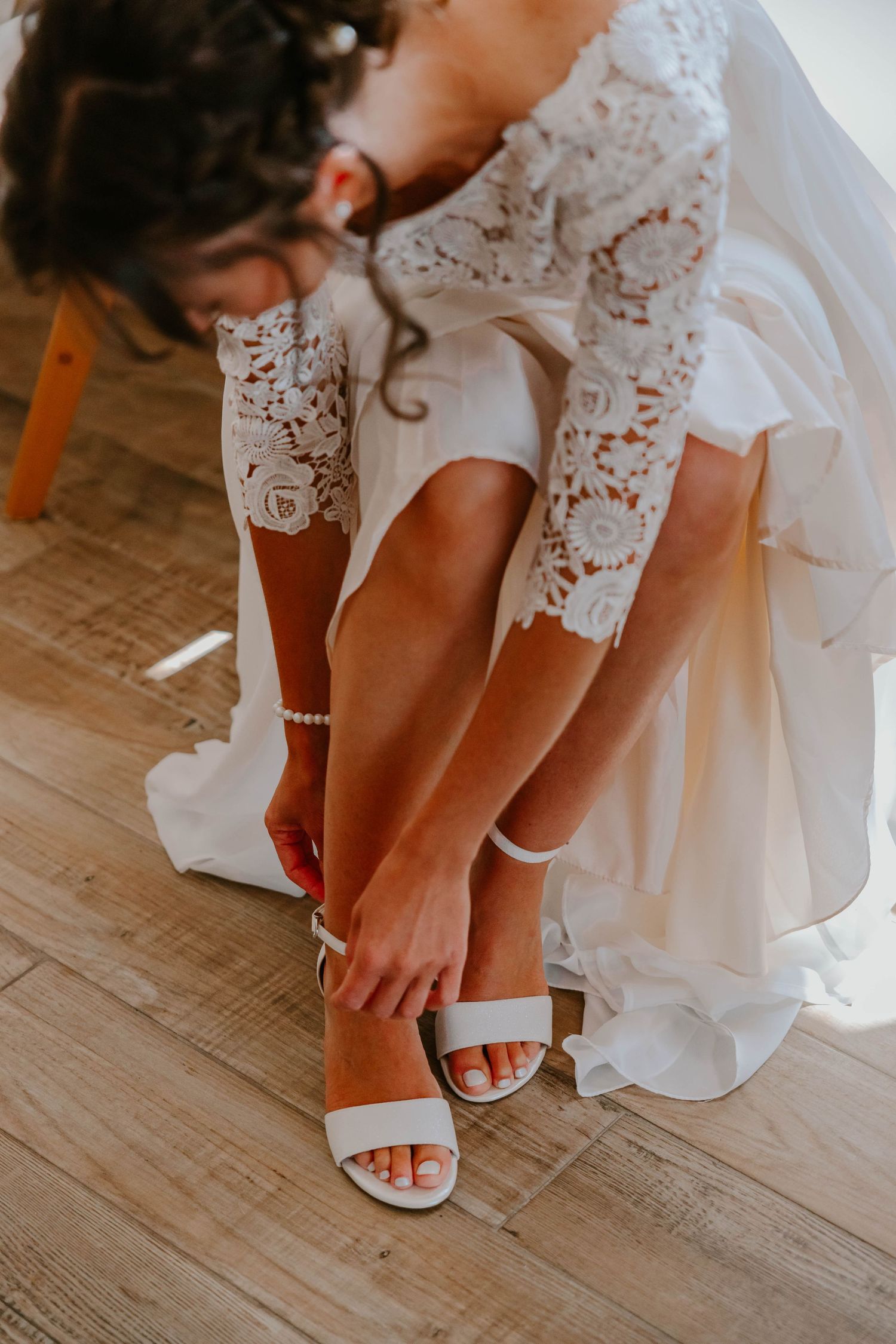 Bride in lace wedding dress fastening white strappy heeled sandals on wooden floor.