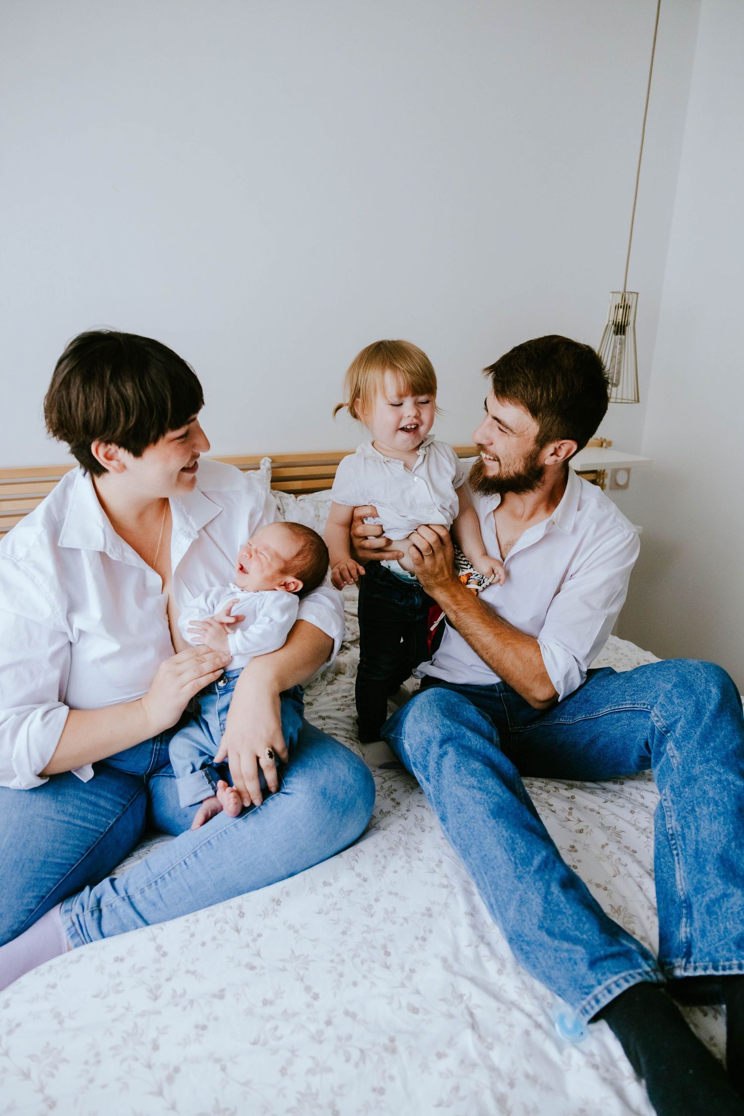 Happy family of four with newborn and laughing toddler enjoying playful moment on bed.