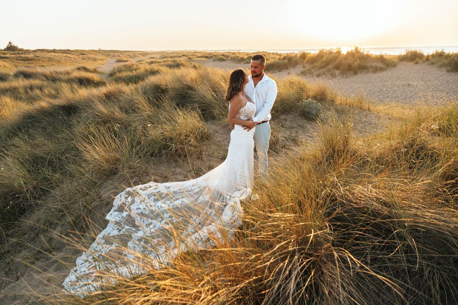 Bride in flowing lace mermaid dress with long train stands with groom amid tall golden beach grass at sunset.