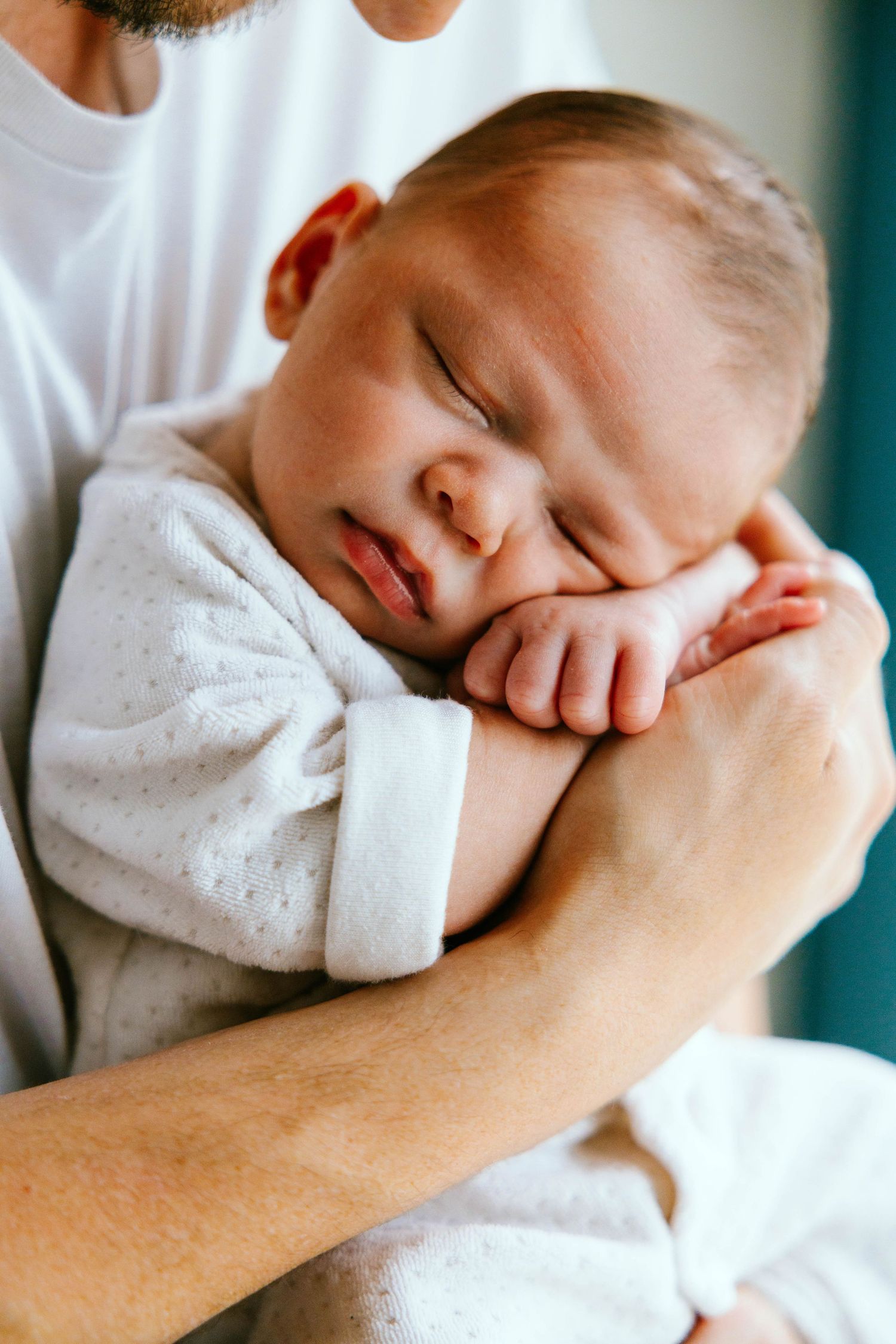 Father gently holding sleeping newborn baby against his chest in white outfit.
