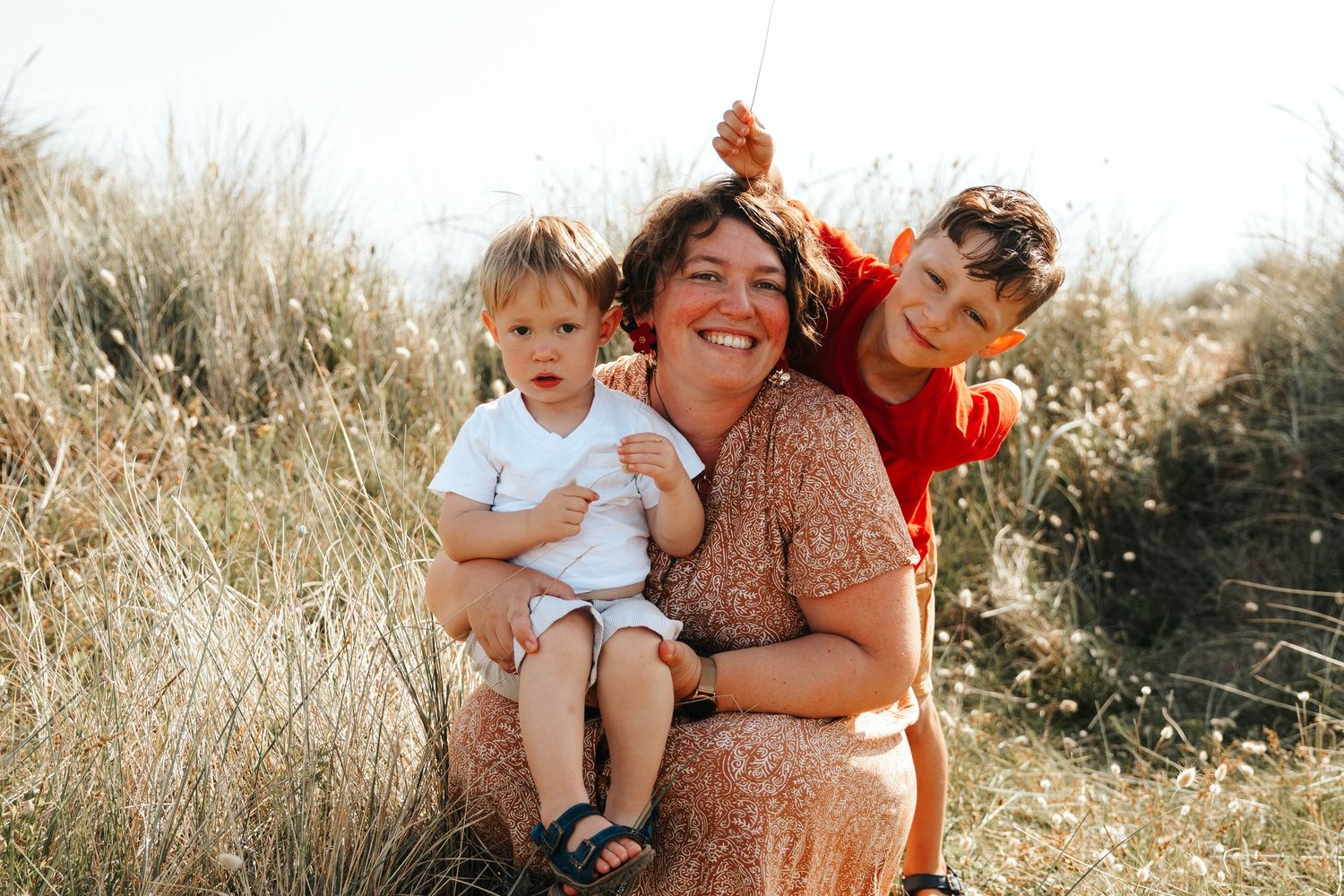 A smiling mother sits in golden grass dunes holding her toddler while her older child hugs her from behind.