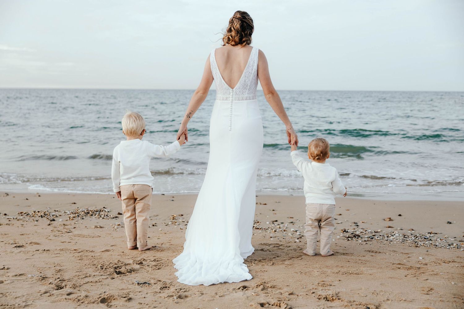 Bride in white wedding gown holds hands with two young children on a sandy beach by the ocean.