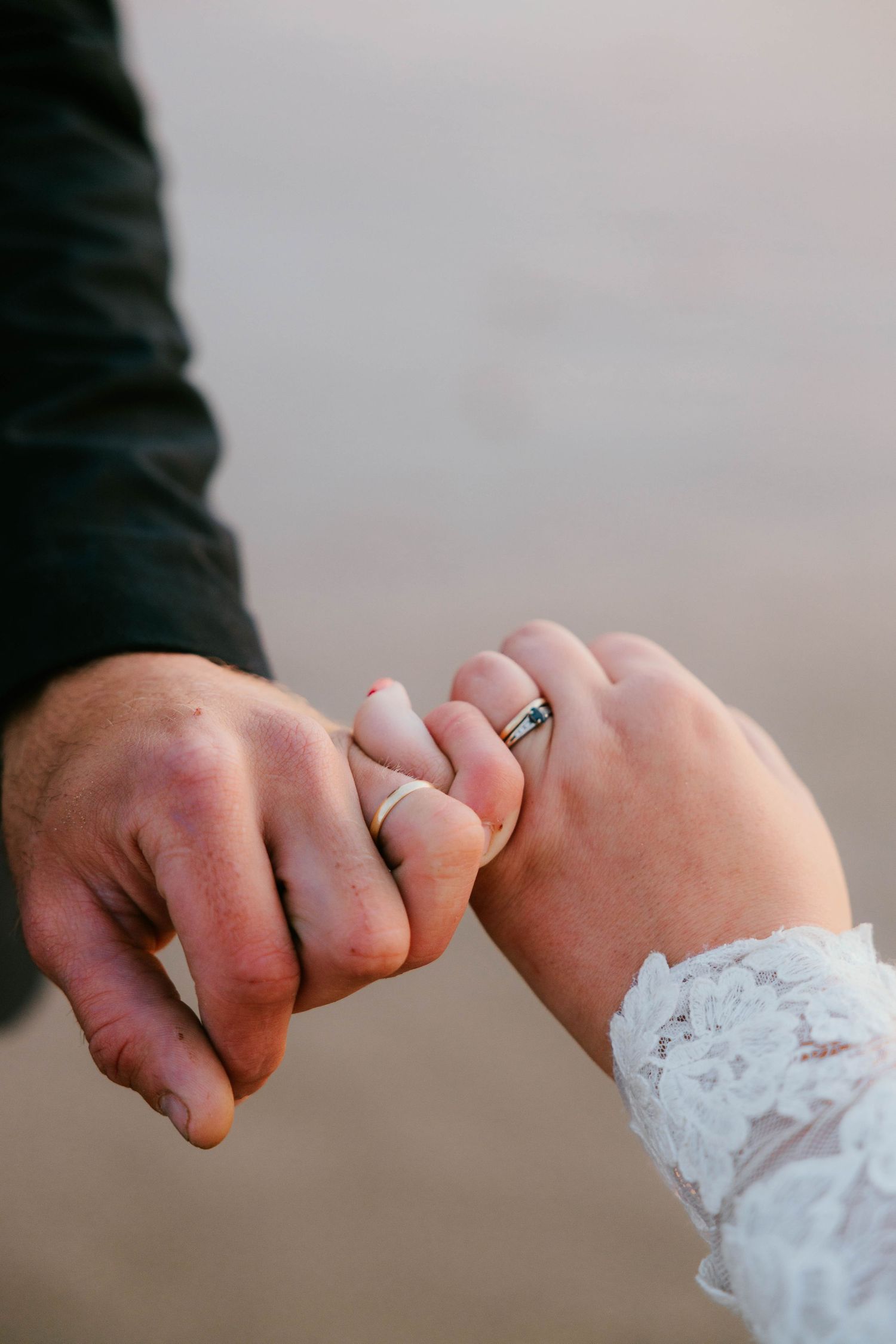 Newlywed couple holding pinkies showing wedding bands, bride in lace sleeve, groom in dark suit, romantic beach setting.