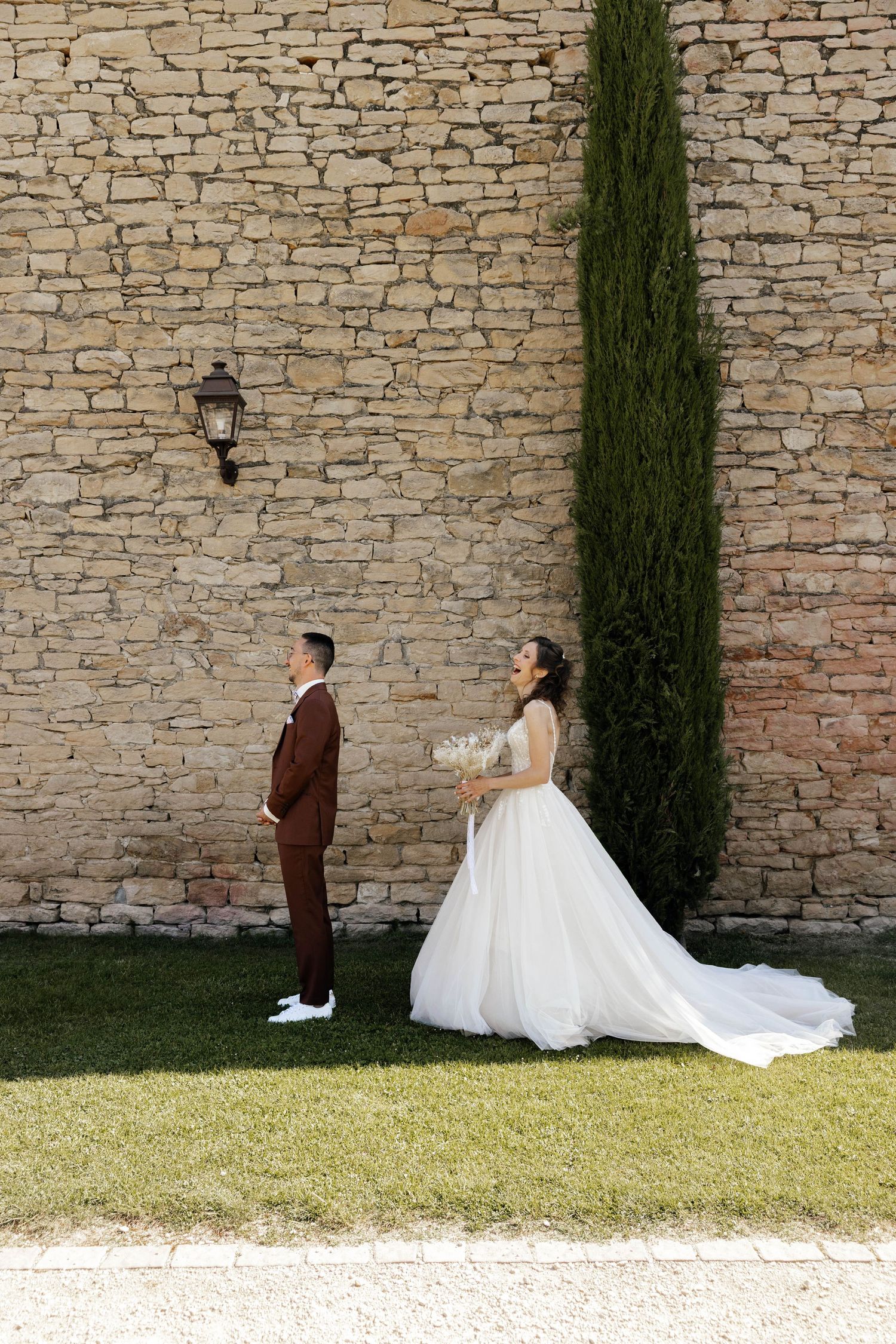 Bride in white gown holding bouquet stands near groom in brown suit against rustic stone wall with cypress tree.