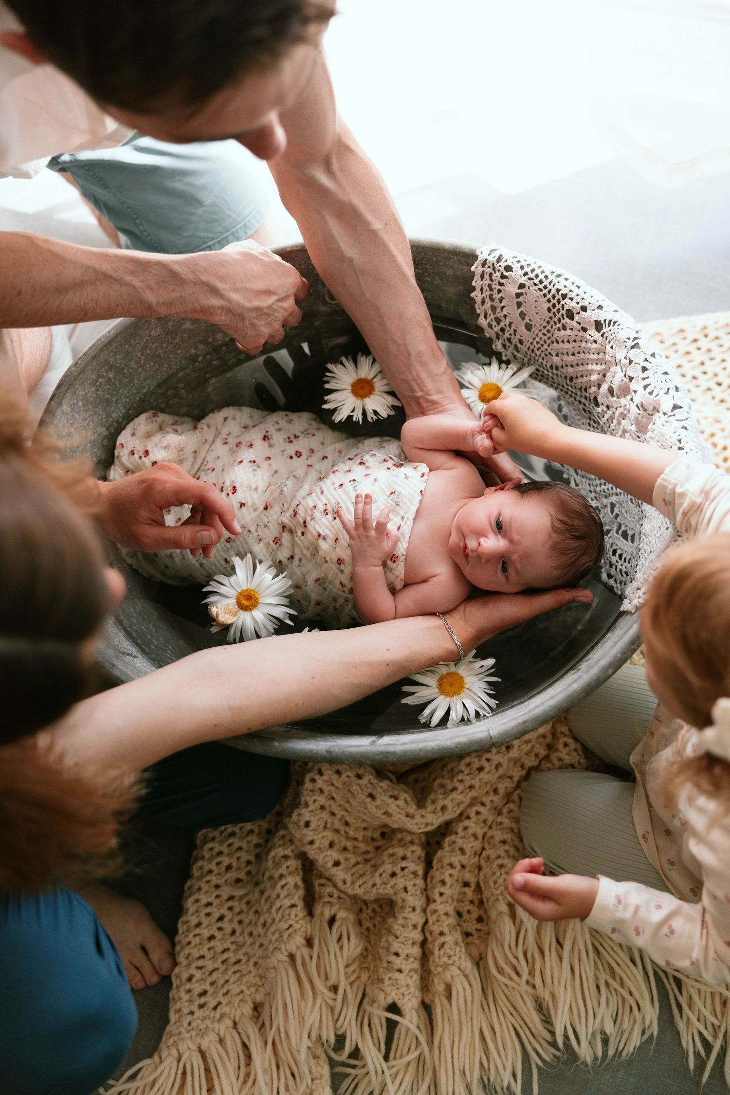 Family with toddler girl surrounds newborn baby lying in rustic bowl decorated with daisy flowers.