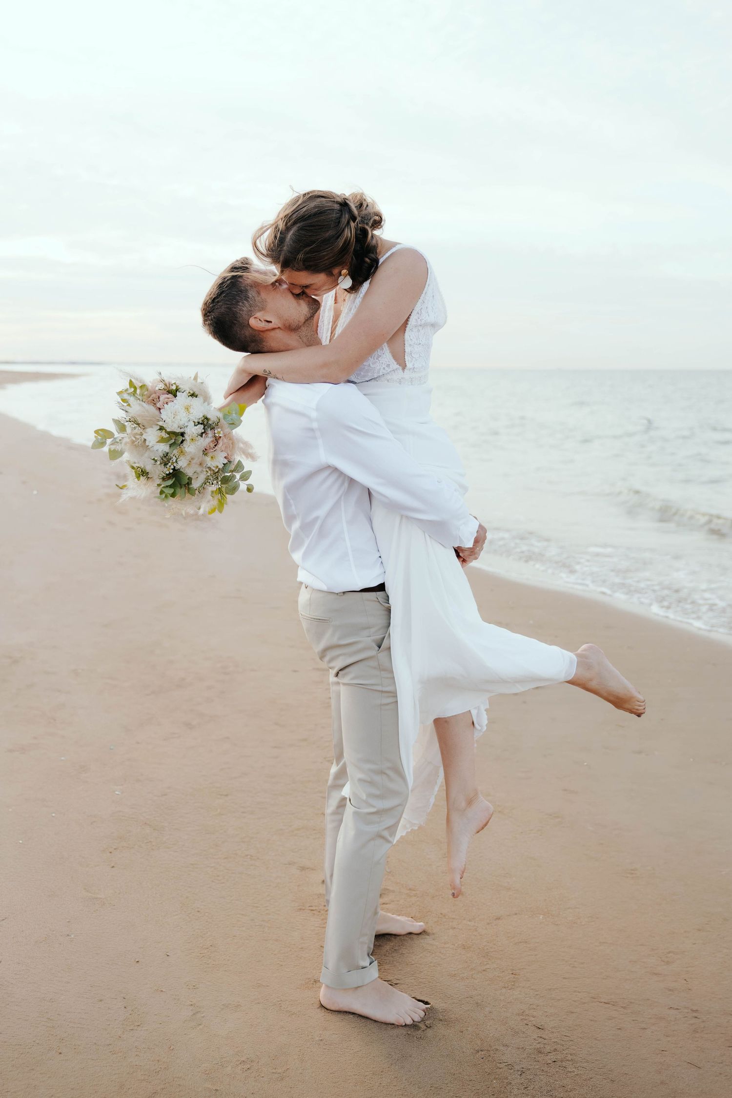 Groom lifting bride for a kiss on the ocean shoreline.
