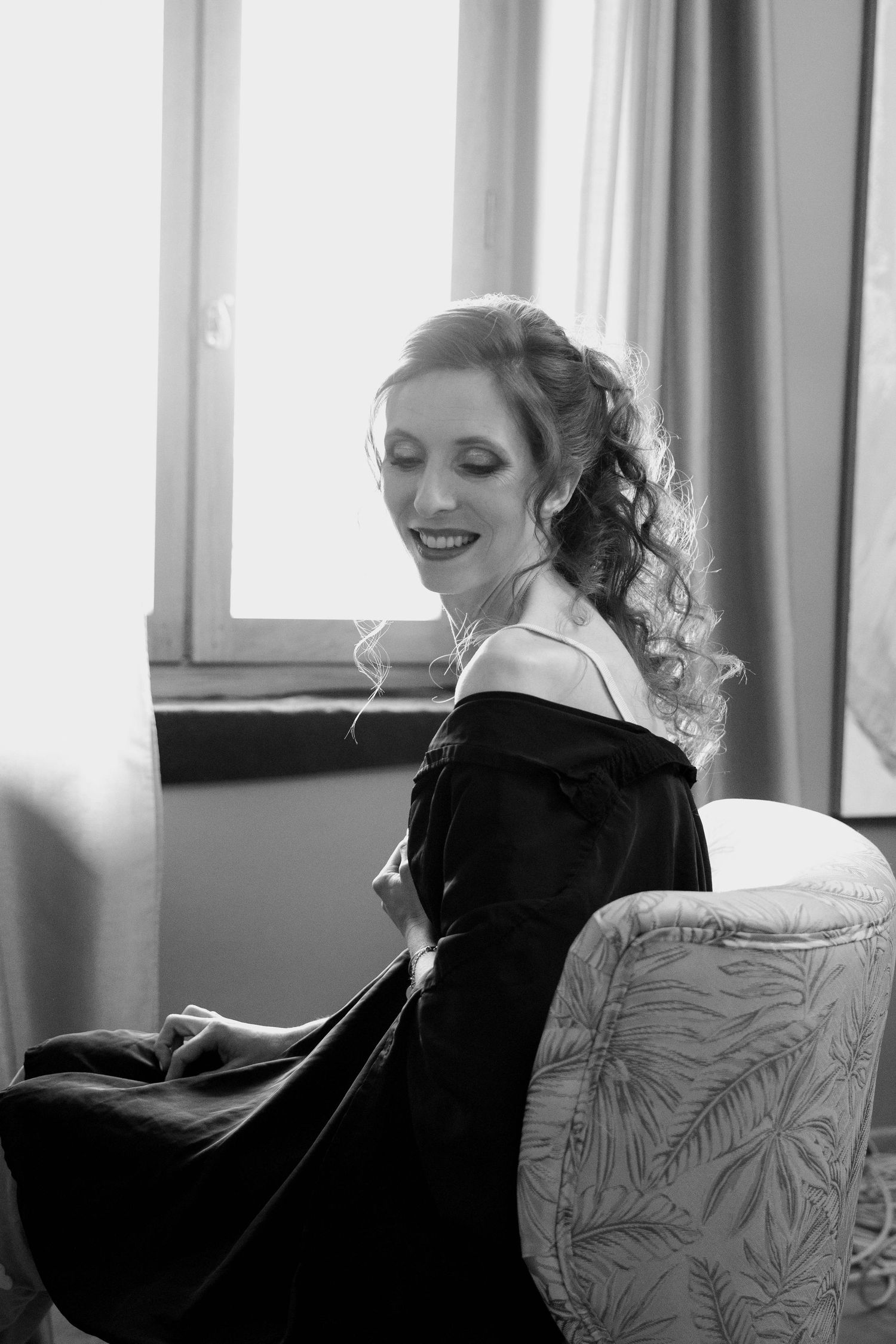Black and white portrait of a smiling bride in an off-shoulder gown seated by a window.