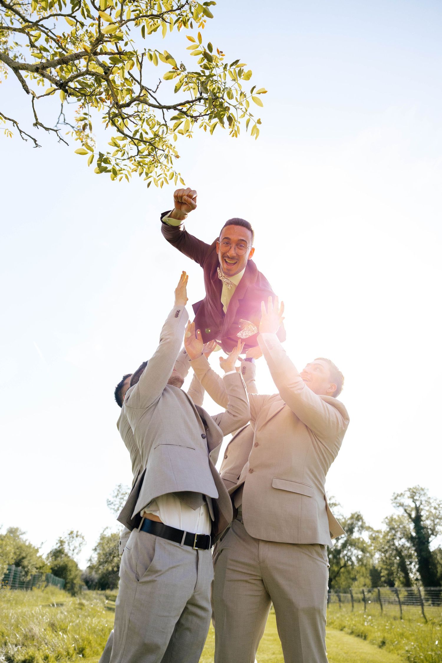 Groomsmen joyfully tossing groom into air outdoors in sunlit garden during wedding celebration.
